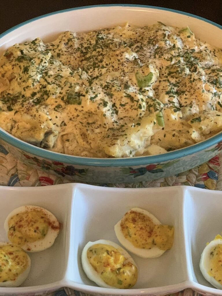 A large dish of creamy potato salad topped with chopped herbs sits behind a tray of deviled eggs, garnished with paprika and herbs, all on a woven placemat.