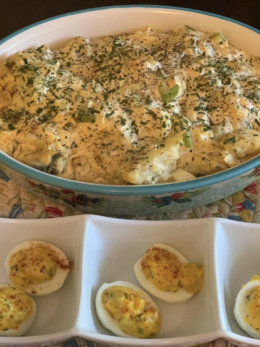 A large dish of creamy potato salad topped with chopped herbs sits behind a tray of deviled eggs, garnished with paprika and herbs, all on a woven placemat.