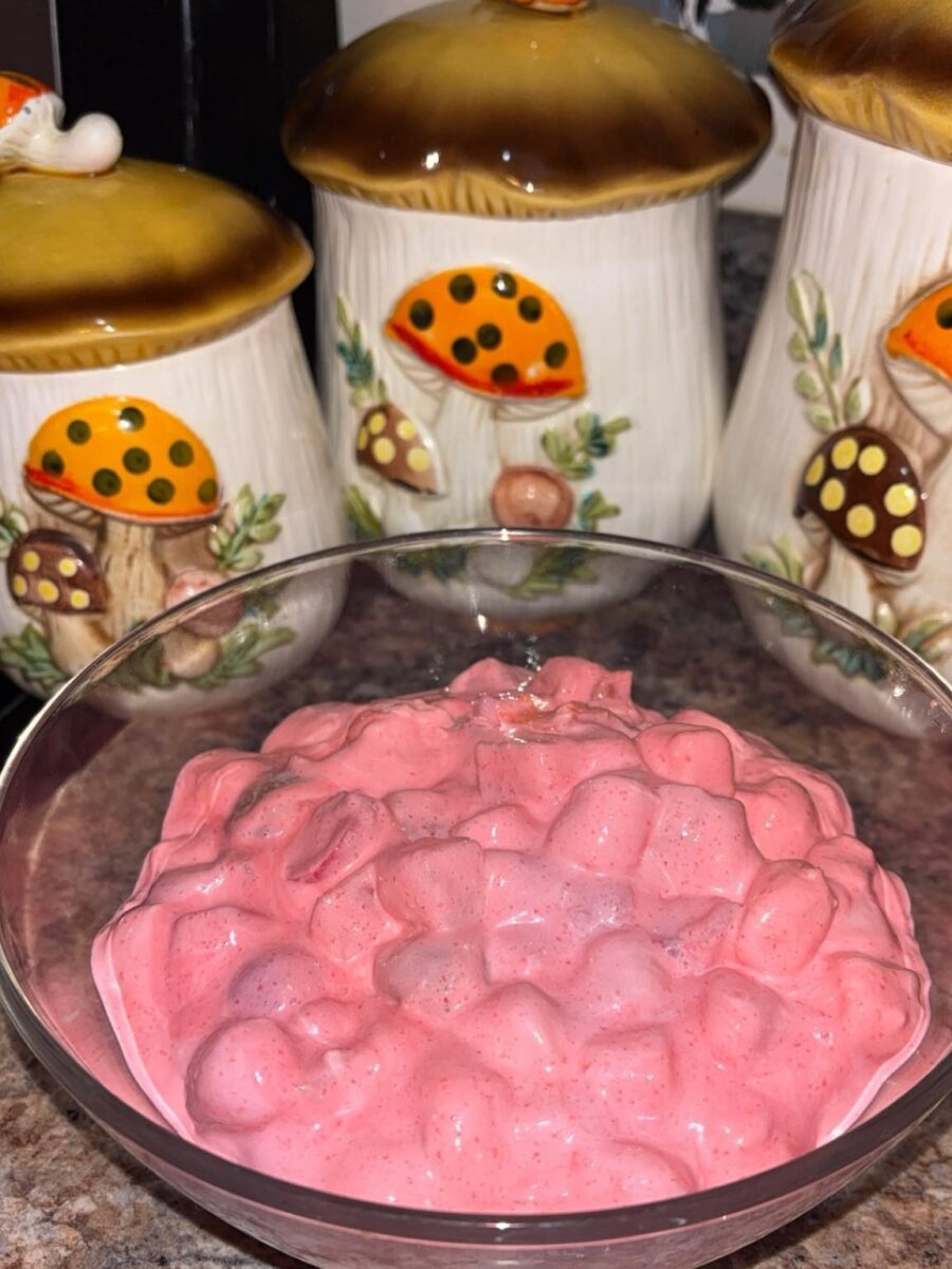 A glass bowl filled with pink, creamy dessert containing mini marshmallows sits on a kitchen counter. In the background, there are three ceramic canisters decorated with colorful mushrooms.