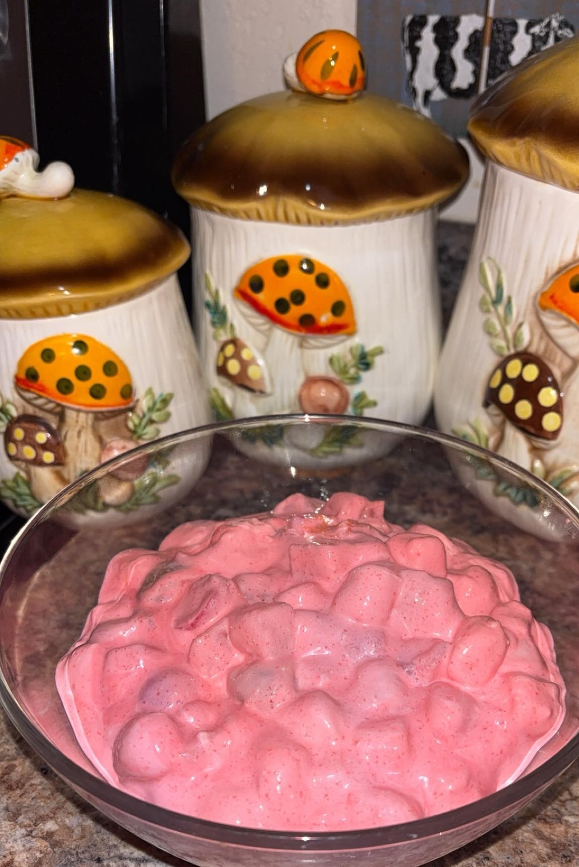 A glass bowl filled with pink, creamy dessert containing marshmallows sits on a countertop in front of three ceramic canisters decorated with colorful mushrooms.