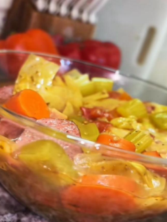 A clear glass bowl filled with a colorful stew containing sliced carrots, celery, sausage, and cabbage, placed on a kitchen counter with tomatoes and kitchen utensils visible in the background.