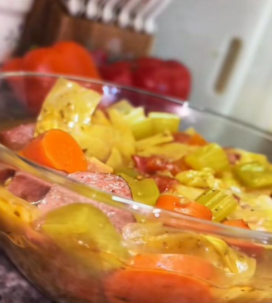 A clear glass bowl filled with a colorful stew containing sliced carrots, celery, sausage, and other vegetables, with tomatoes and kitchen utensils blurred in the background.