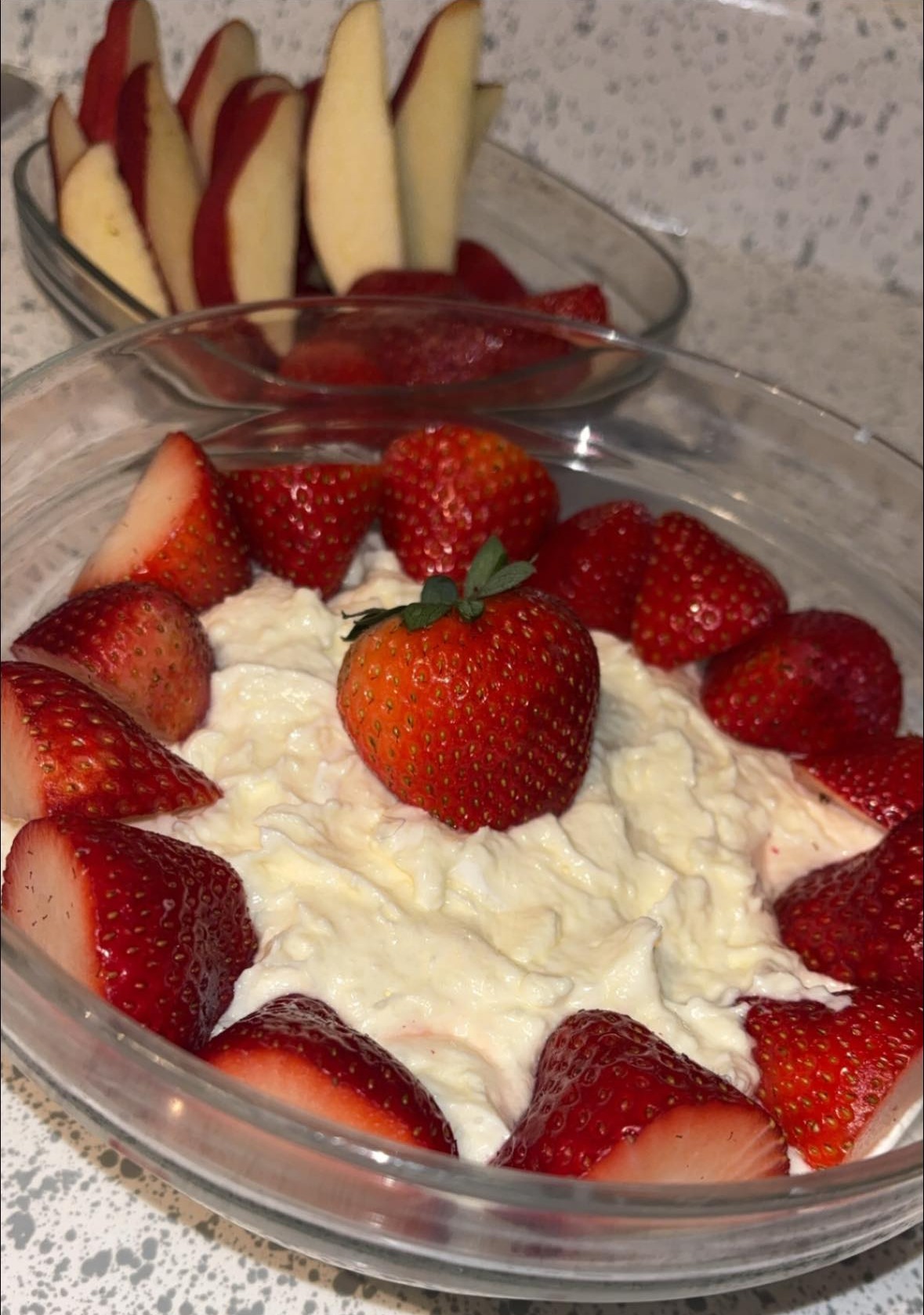 A glass bowl filled with creamy dip topped with a whole strawberry and surrounded by halved strawberries. In the background, another bowl contains sliced red apples arranged upright. The setting is on a speckled countertop.