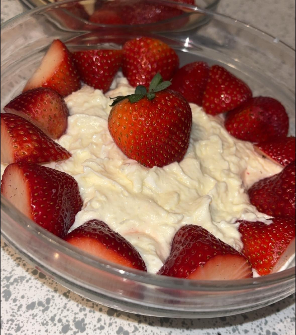 A glass bowl filled with creamy rice pudding, topped with a whole strawberry in the center and surrounded by halved strawberries, placed on a speckled countertop.