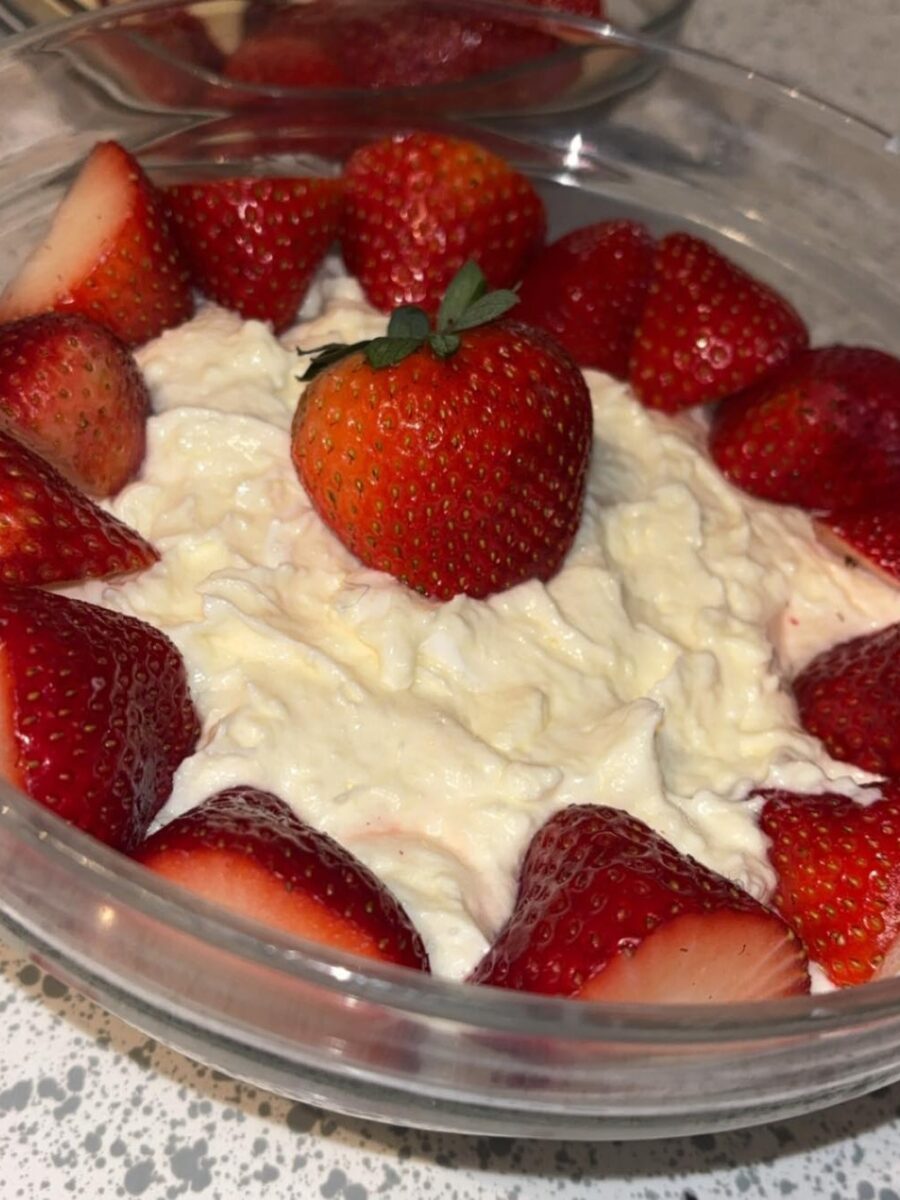 A bowl of creamy dessert topped with whole and halved fresh strawberries, arranged around the edge and one placed in the center. The bowl is on a speckled countertop.