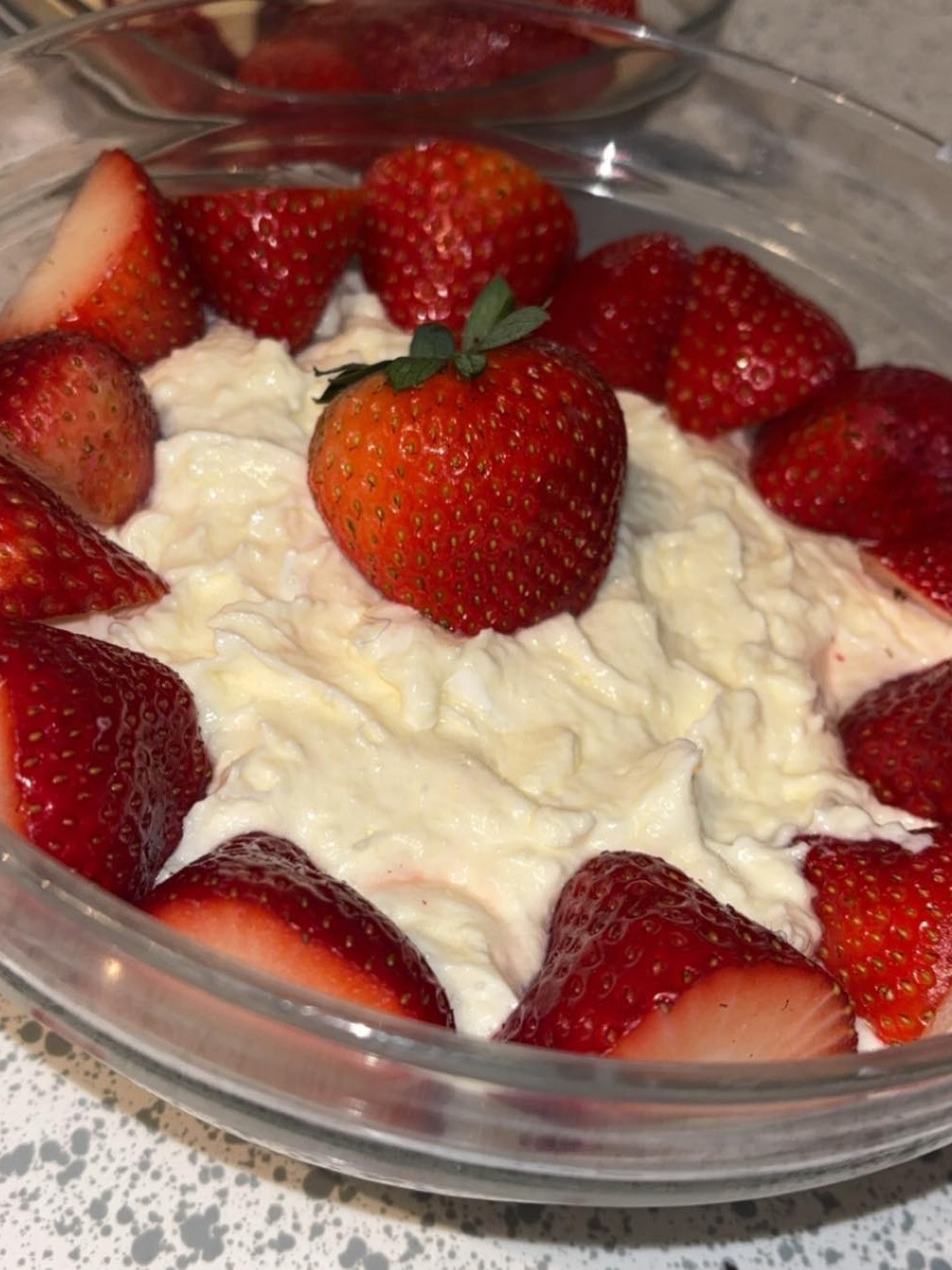 A bowl of creamy dessert topped with whole and halved fresh strawberries, arranged around the edge and one placed in the center. The bowl is on a speckled countertop.