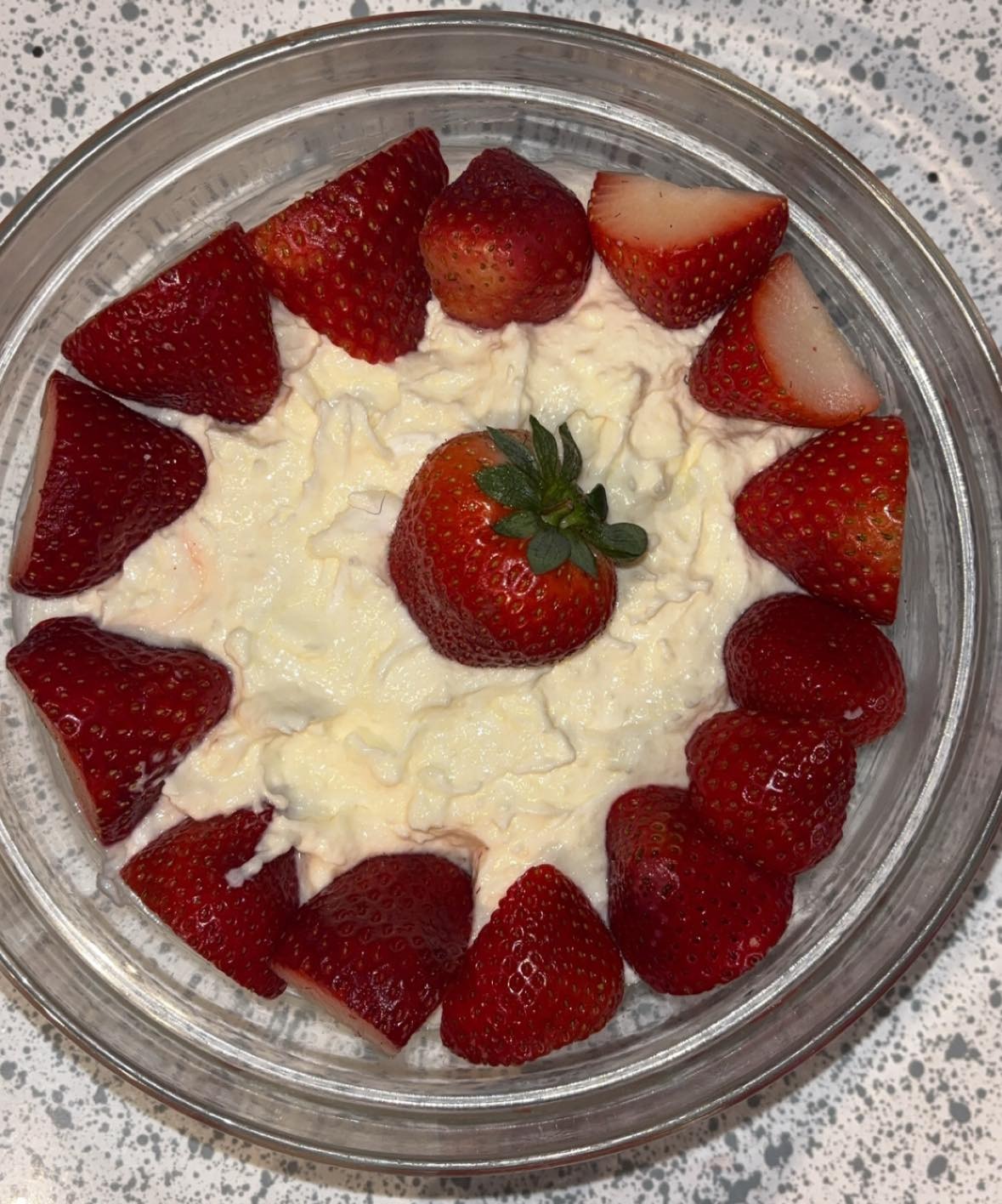 A glass bowl filled with creamy rice pudding, topped with a ring of halved strawberries around the edge and a whole strawberry placed in the center. The bowl is on a speckled countertop.