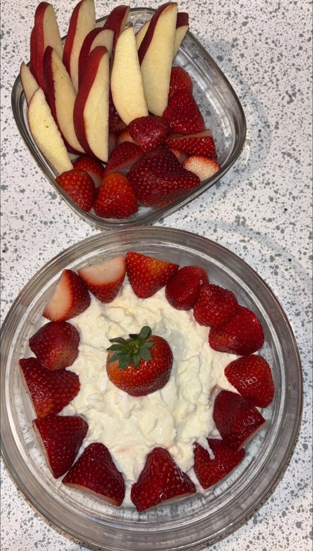 Two glass bowls on a speckled countertop: one filled with red apple slices and whole strawberries, the other with strawberries arranged around a creamy dip, with a whole strawberry in the center.