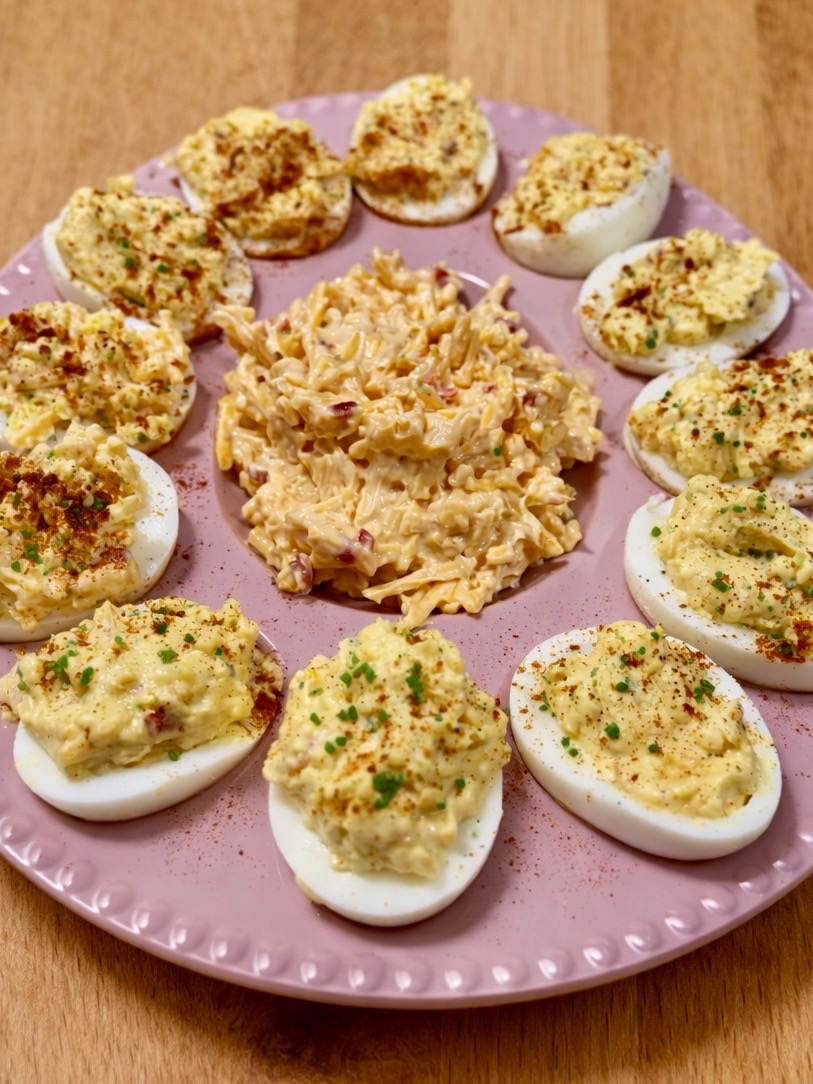 A pink plate holds ten deviled eggs sprinkled with herbs and paprika, arranged around a mound of creamy pimento cheese spread. The plate sits on a wooden surface.