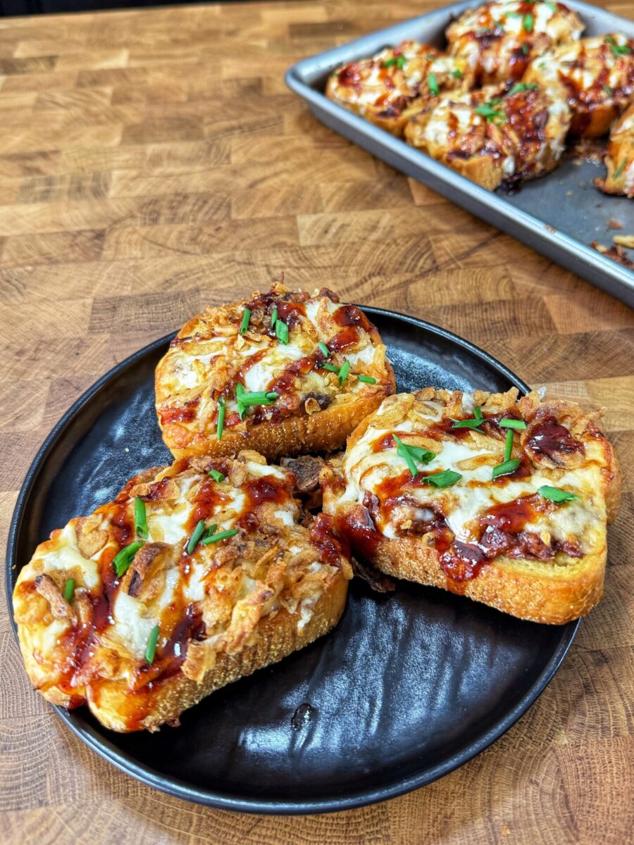 Three pieces of cheesy barbecue chicken toast garnished with chopped chives sit on a black plate, with more pieces on a baking tray in the background on a wooden surface.