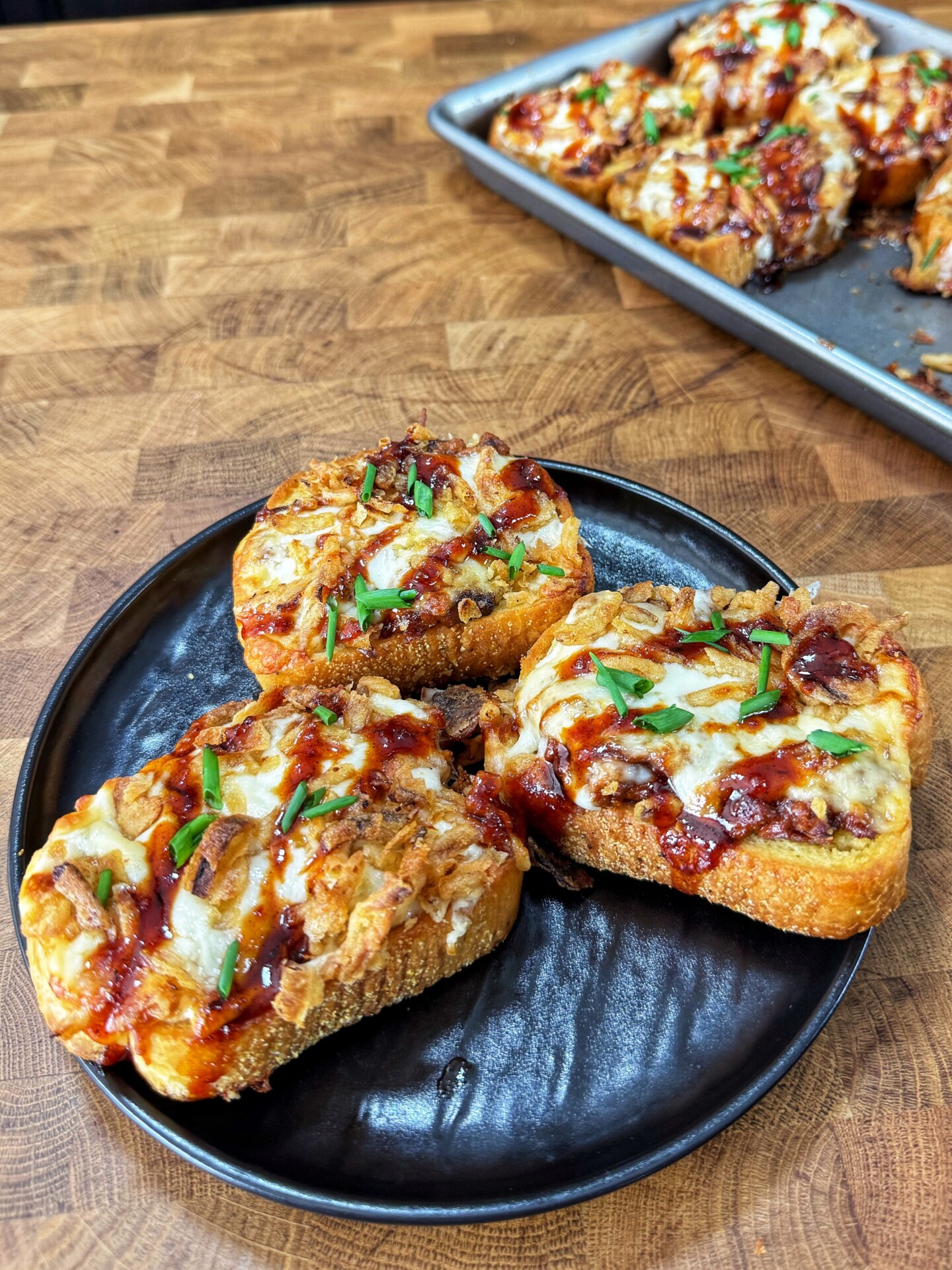 Three pieces of cheesy barbecue chicken toast garnished with chopped chives sit on a black plate, with more pieces on a baking tray in the background on a wooden surface.