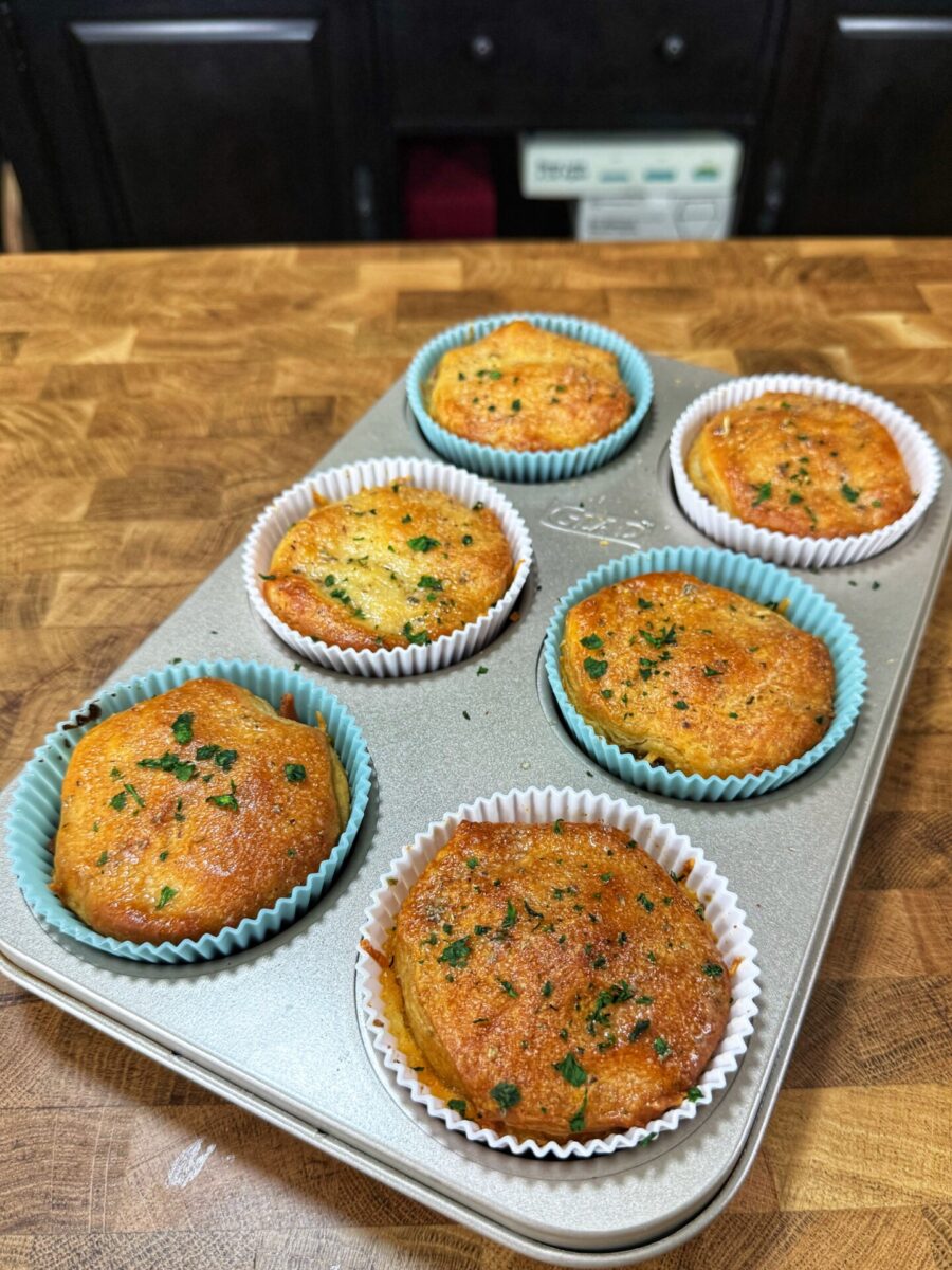 Six golden-brown muffins with herbs on top sit in a muffin tin lined with colorful paper cups, placed on a wooden countertop. The background shows dark kitchen cabinets.