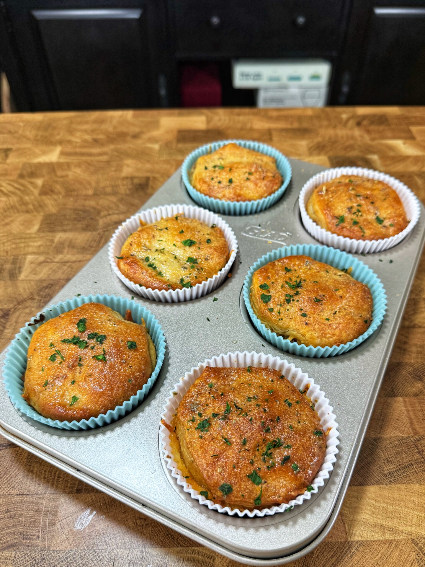Six golden-brown muffins with herbs on top sit in a muffin tin lined with colorful paper cups, placed on a wooden countertop. The background shows dark kitchen cabinets.