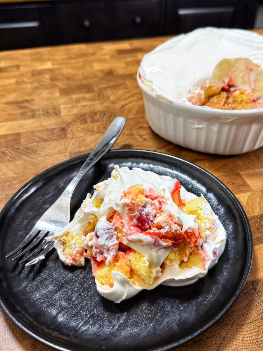 A slice of strawberry shortcake with whipped cream on a black plate, next to a fork. In the background, the partially served dessert is in a white baking dish on a wooden countertop.