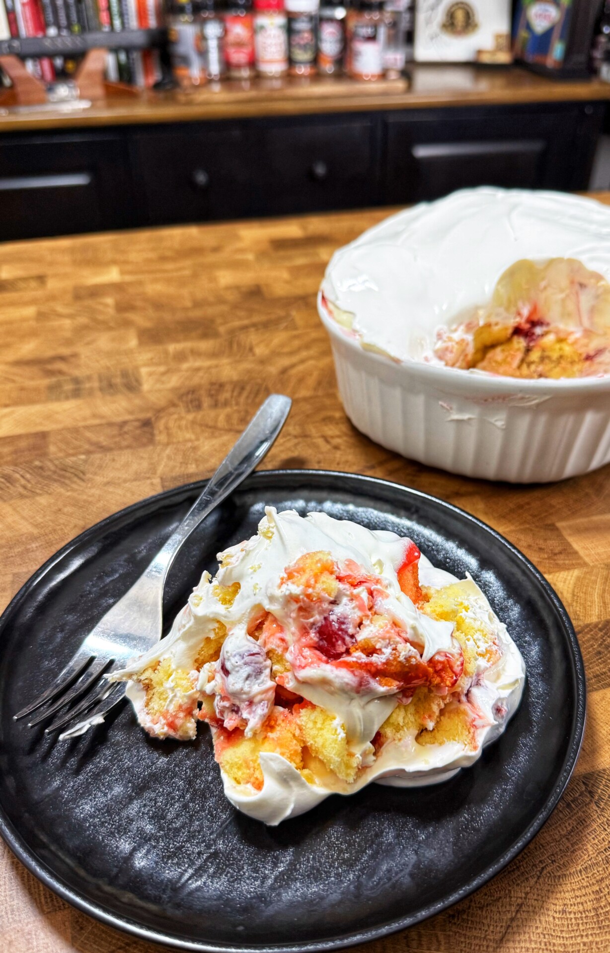 A plate with a serving of dessert made of cake, strawberries, and whipped cream sits on a wooden counter, with a fork beside it. In the background, the rest of the dessert is in a large white dish.