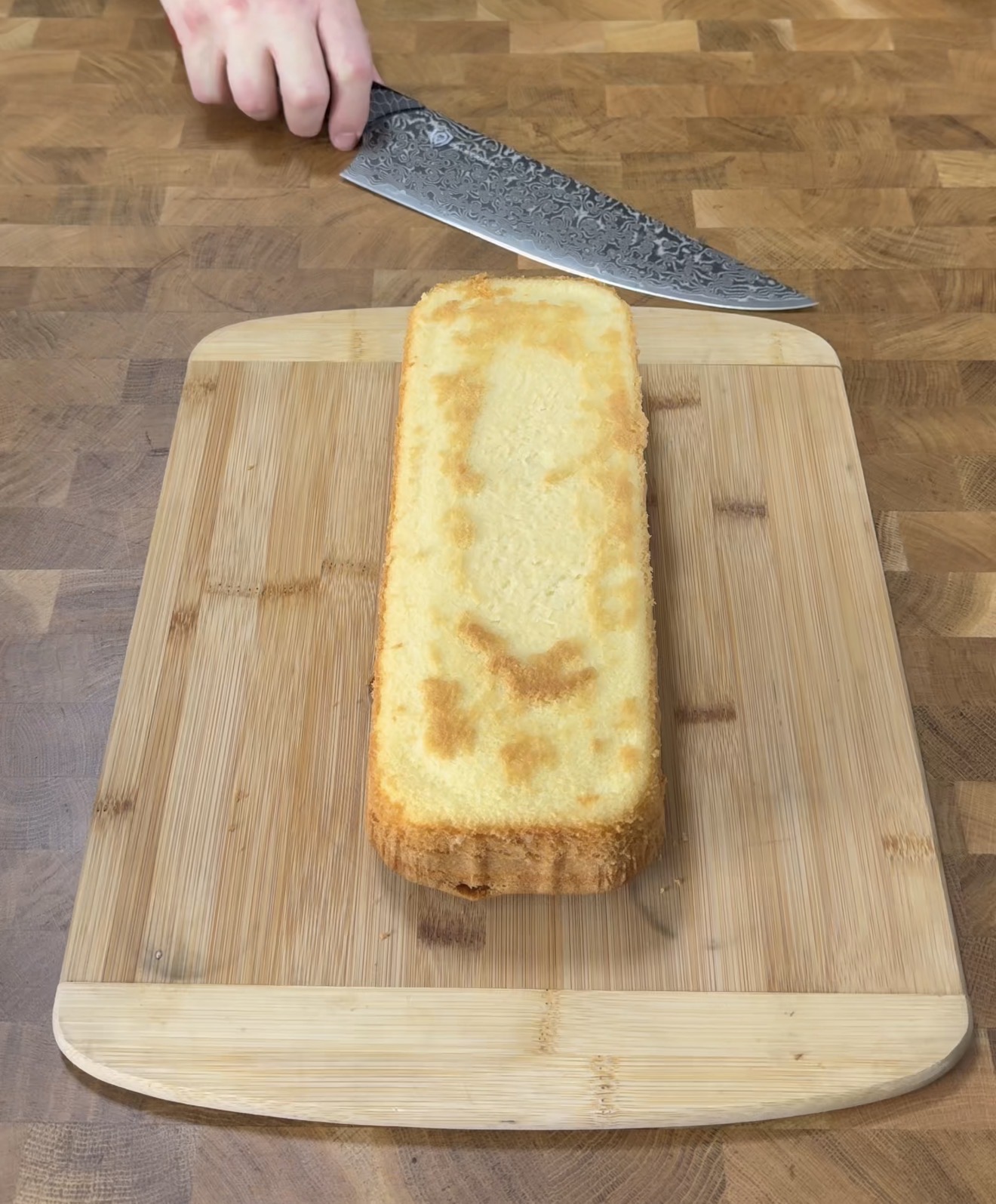 A hand holds a large knife above a rectangular loaf cake on a wooden cutting board, preparing to slice it. The board sits on a wooden countertop.
