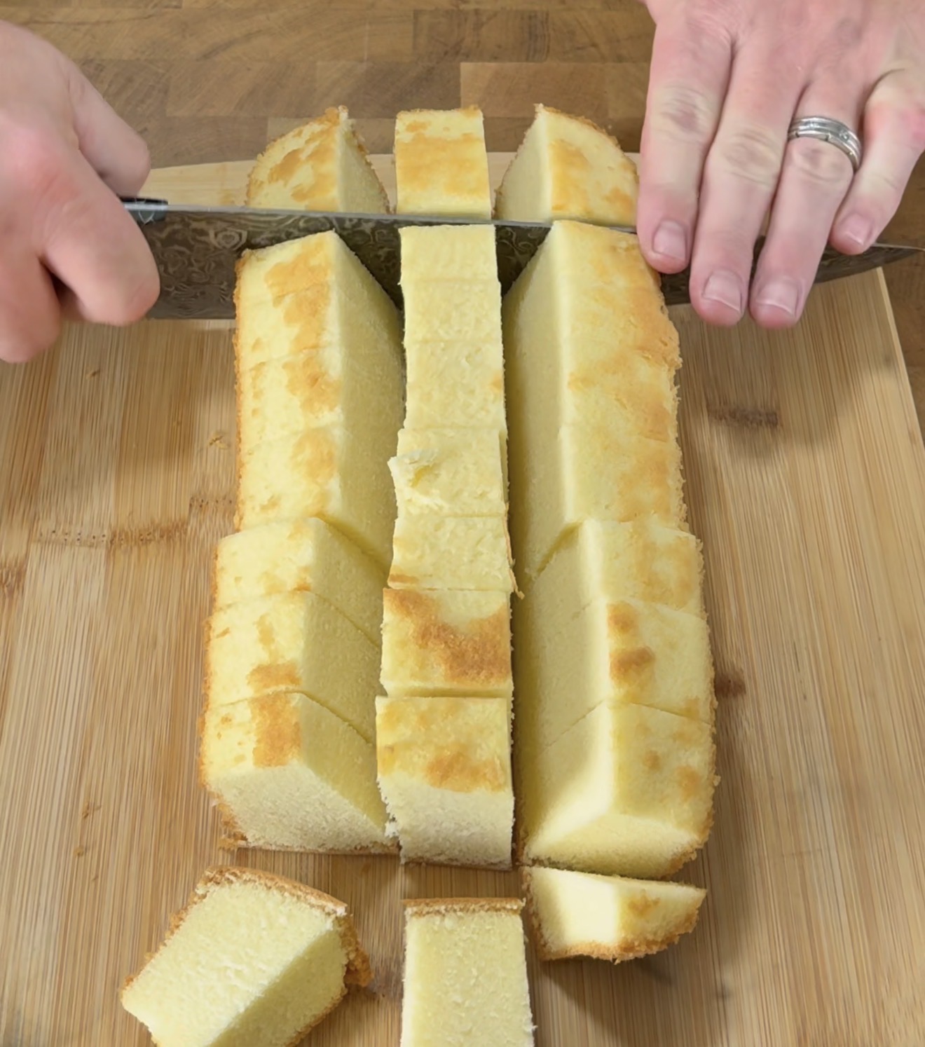 A person slices a rectangular loaf of pound cake into even cubes on a wooden cutting board, using a large knife.