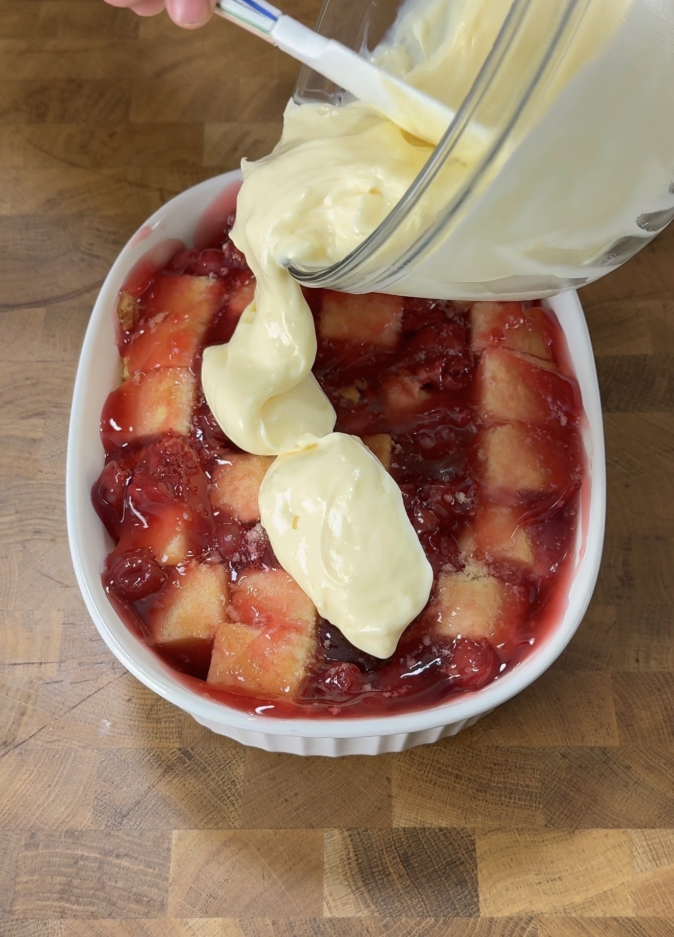 A glass bowl pours creamy yellow pudding over a cherry dessert with a checkerboard pattern in a white oval dish on a wooden surface.