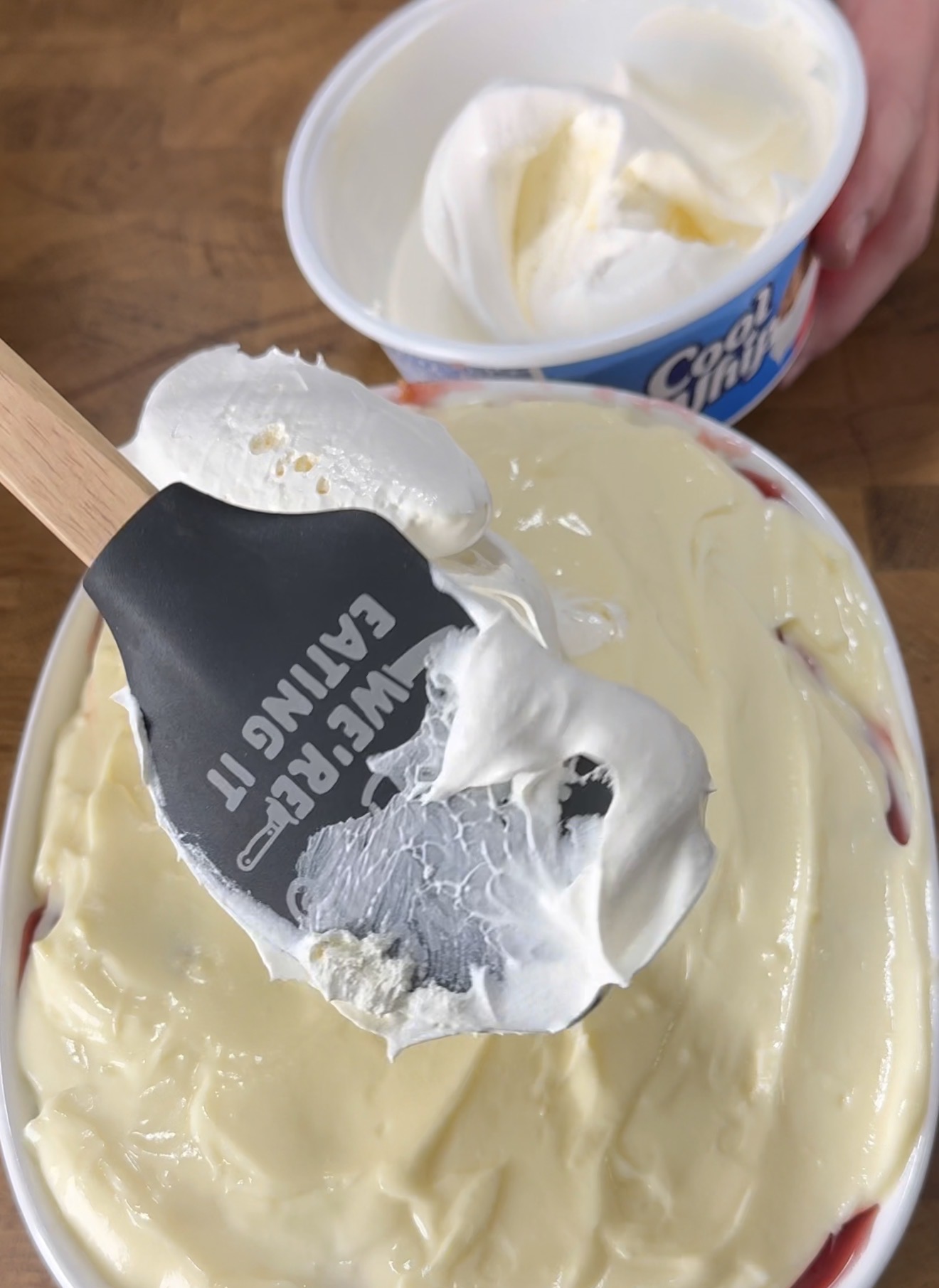 A spatula with “I’D RATHER BE EATING IT” printed on it spreads whipped topping over a creamy dessert in a dish. An open tub of Cool Whip is in the background on a wooden surface.