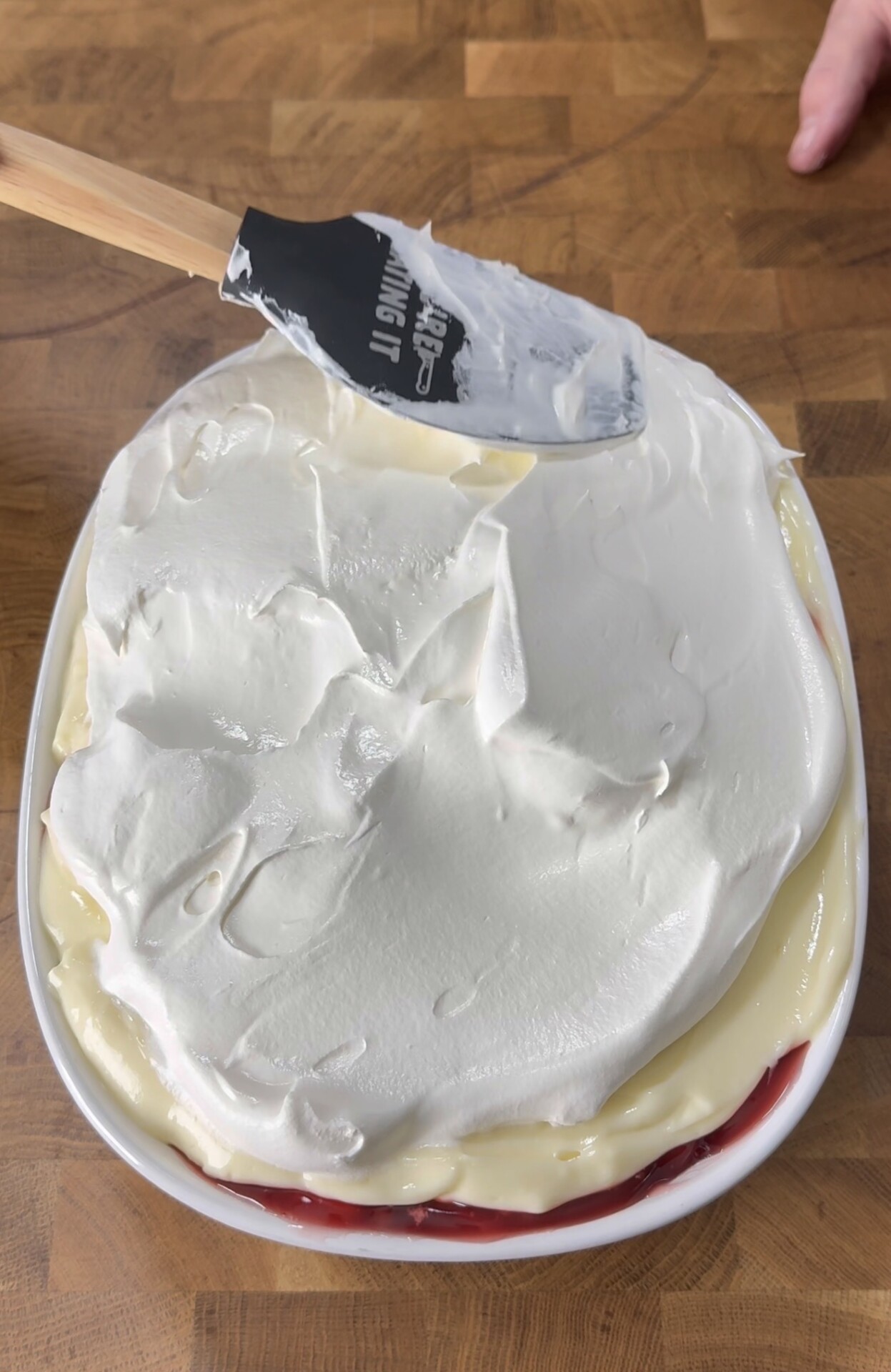 A hand uses a black spatula to spread a thick layer of whipped cream over a dessert with visible custard and fruit layers in a white oval dish on a wooden surface.
