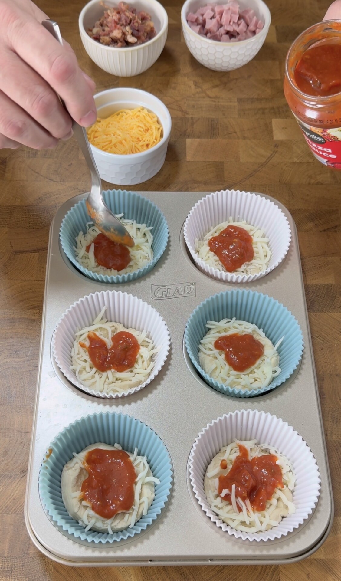 A hand spooning tomato sauce onto cheese-topped dough in cupcake liners in a muffin tray. Bowls of shredded cheese, diced ham, and bacon bits are on the wooden table.