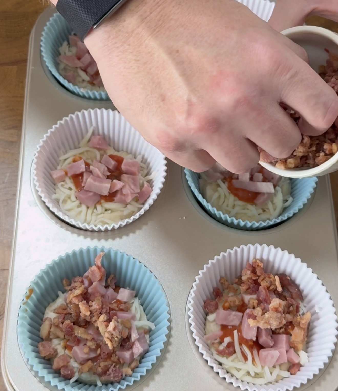 A person sprinkles chopped bacon into blue and white muffin liners filled with shredded cheese, ham, and sauce, preparing a savory dish in a muffin tin.