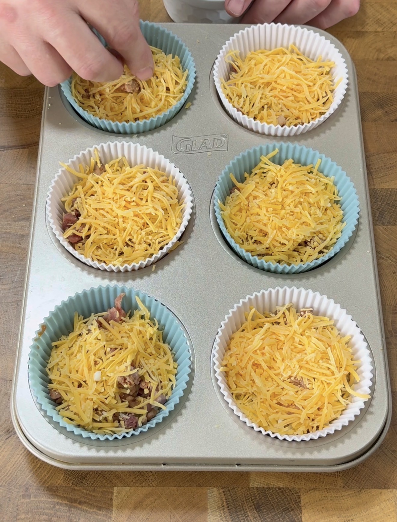 A hand sprinkles shredded cheese into silicone muffin cups in a muffin tin, with each cup filled with cheese and visible bits of cooked bacon. The tin sits on a wooden surface.