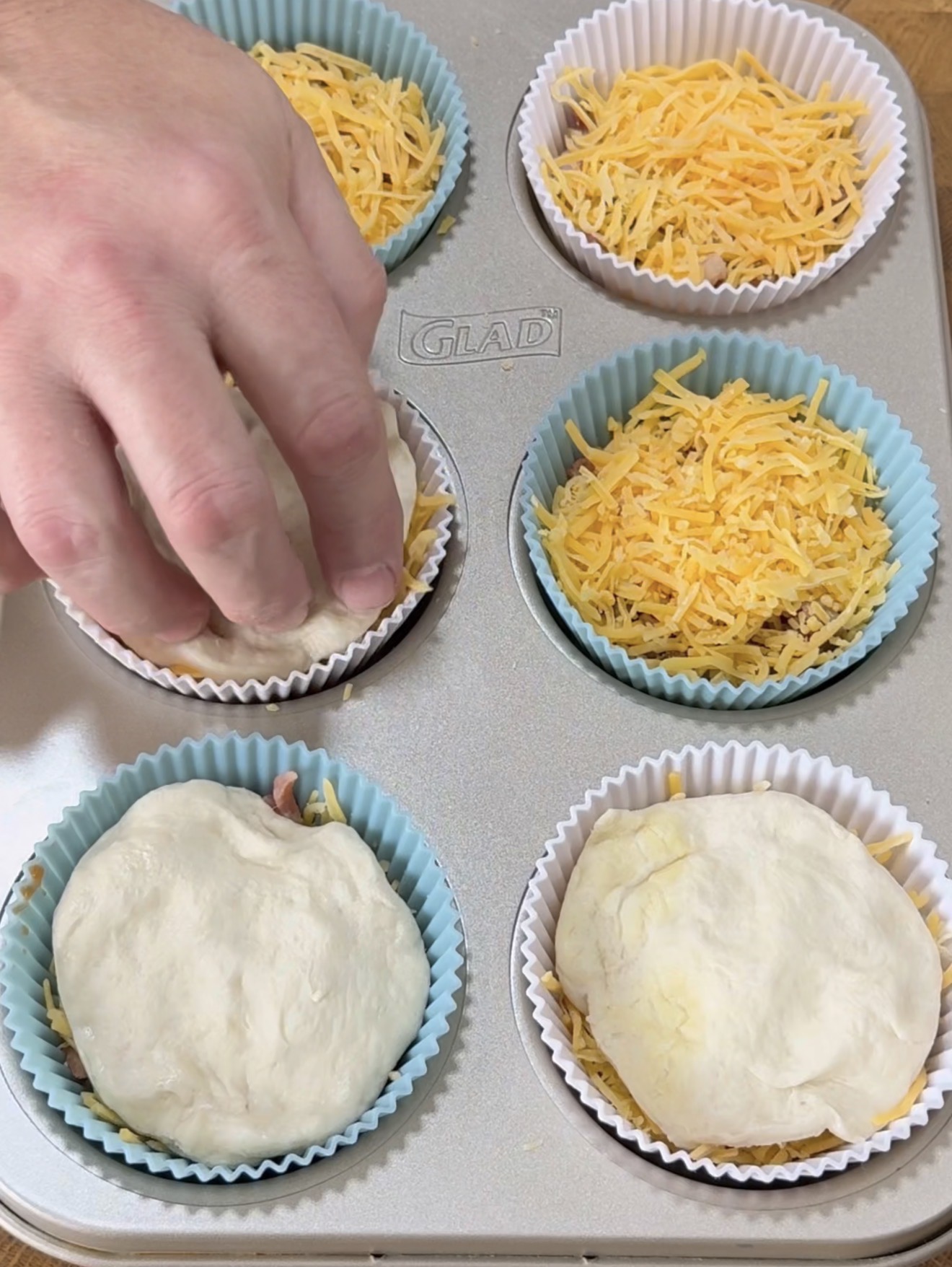 A hand places dough over filling in a muffin tin lined with silicone cups. Some cups are already covered with dough, while others are filled with shredded cheese.