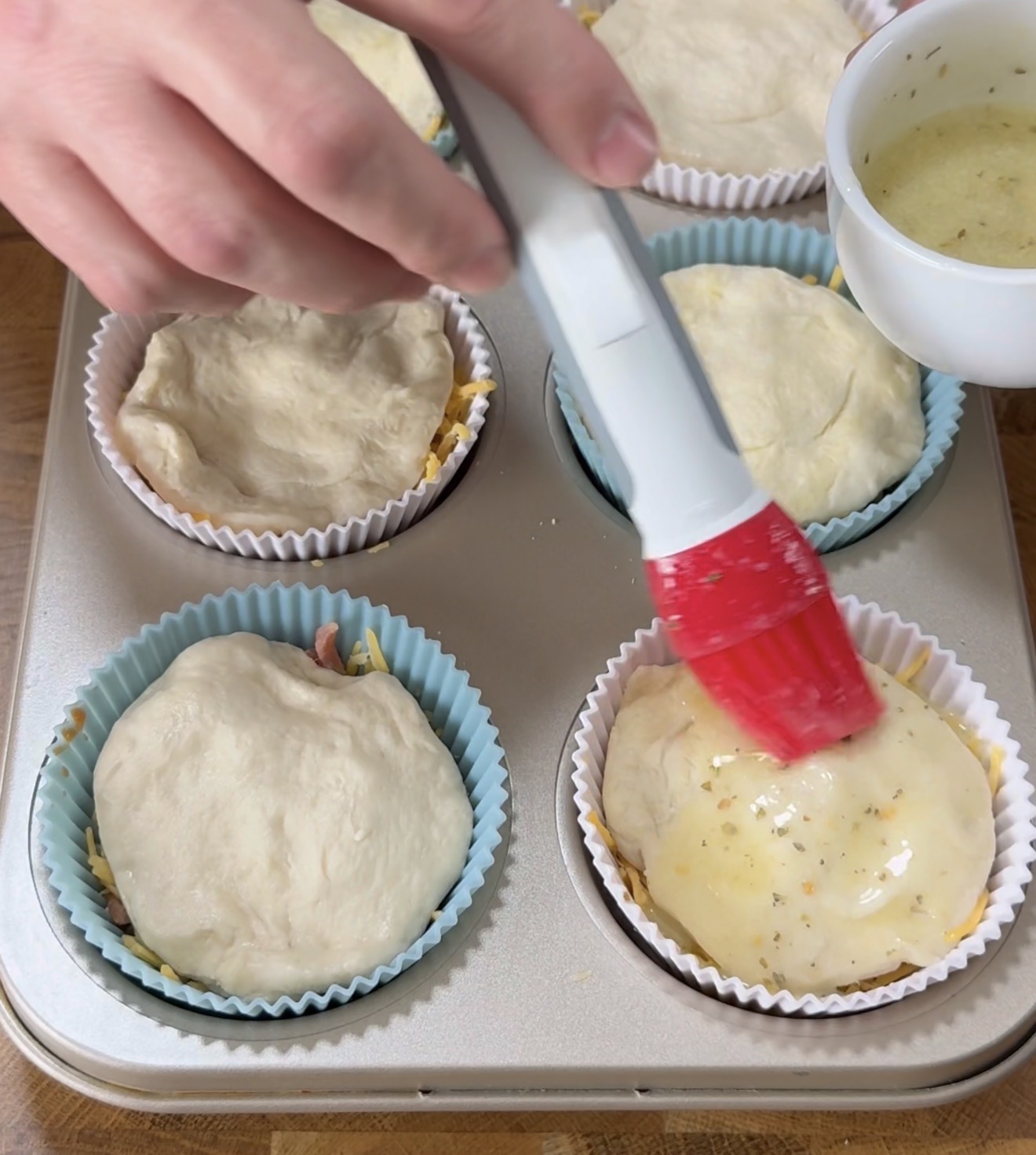 A hand uses a red pastry brush to apply a liquid glaze onto dough-topped muffin cups in a muffin tin. The dough covers fillings in silicone baking cups, and a bowl of glaze sits nearby.