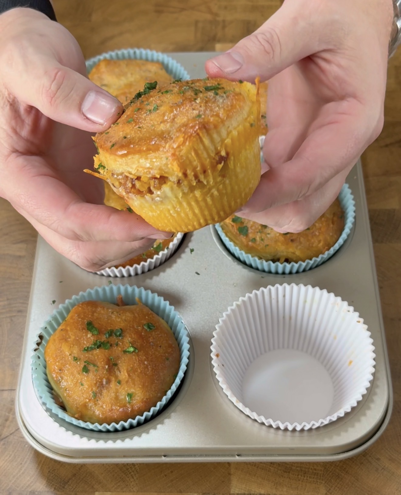 A person holds a savory muffin topped with herbs above a muffin tin containing more muffins, some in blue and white paper liners, on a wooden surface.
