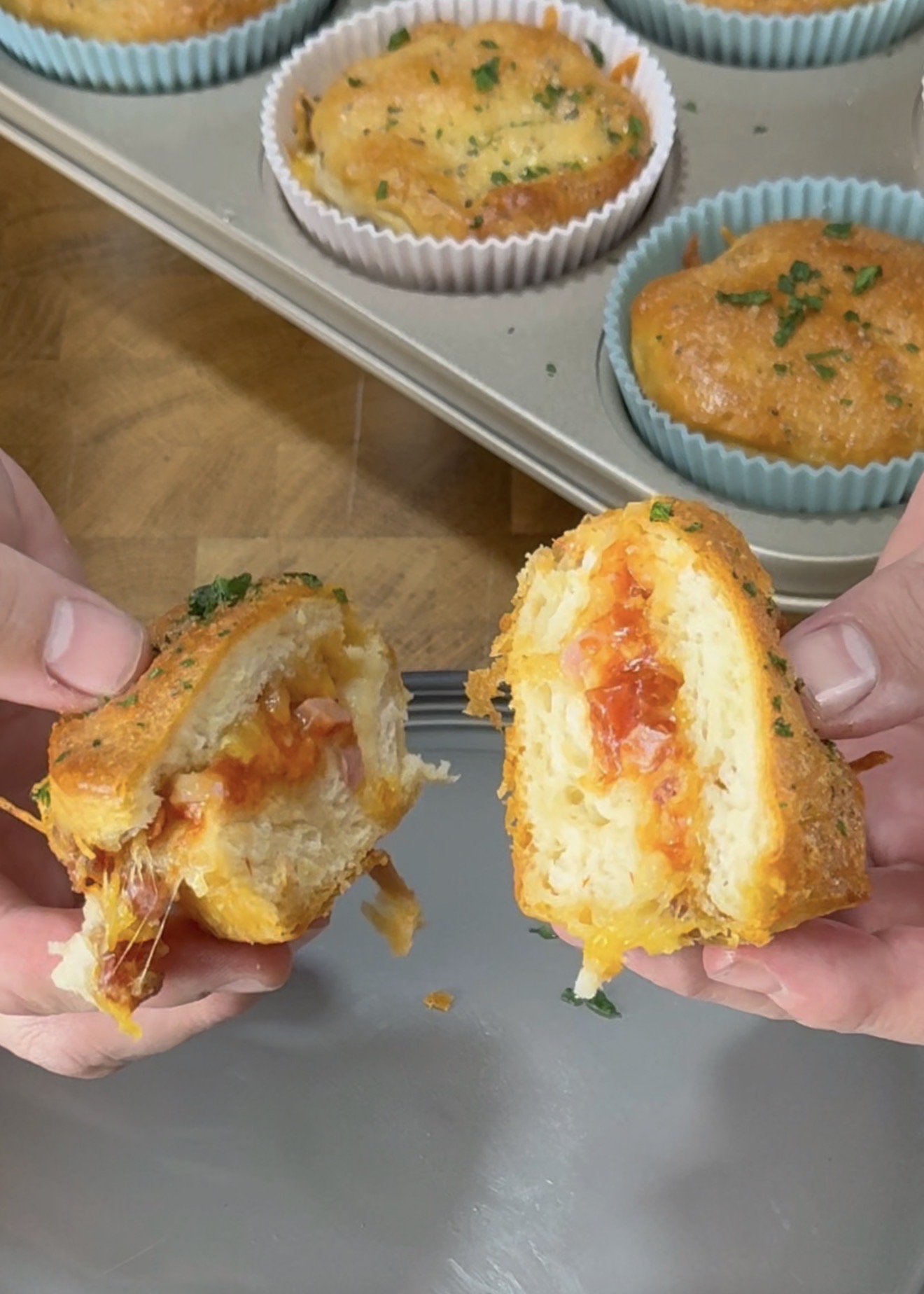 Hands holding and pulling apart a cheesy biscuit muffin, revealing a gooey tomato and cheese filling. A muffin tin with more muffins in colorful liners is in the background on a wooden surface.