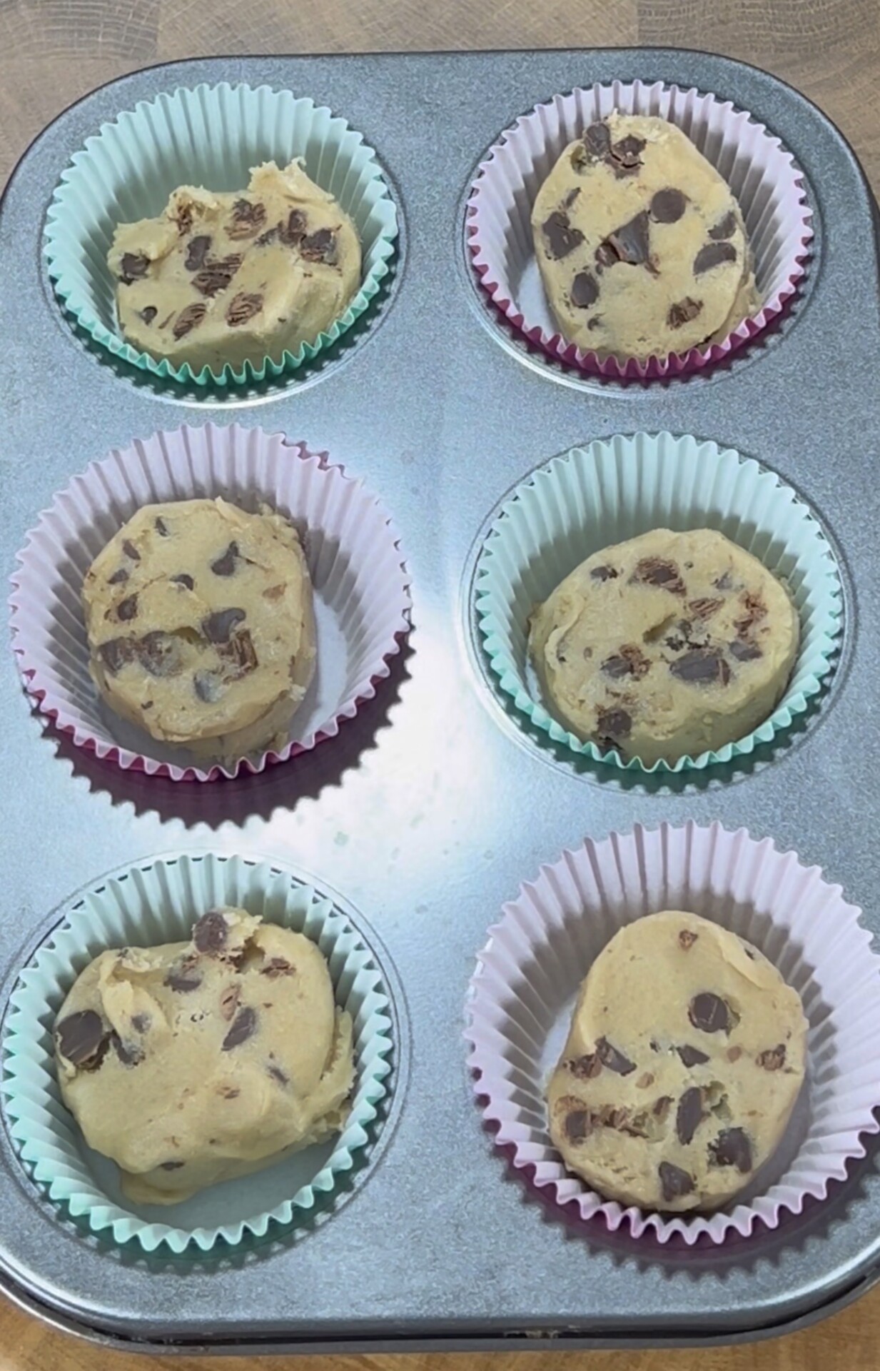 Six muffin tin cups, each lined with colorful paper liners, are filled with raw chocolate chip cookie dough, ready to be baked.
