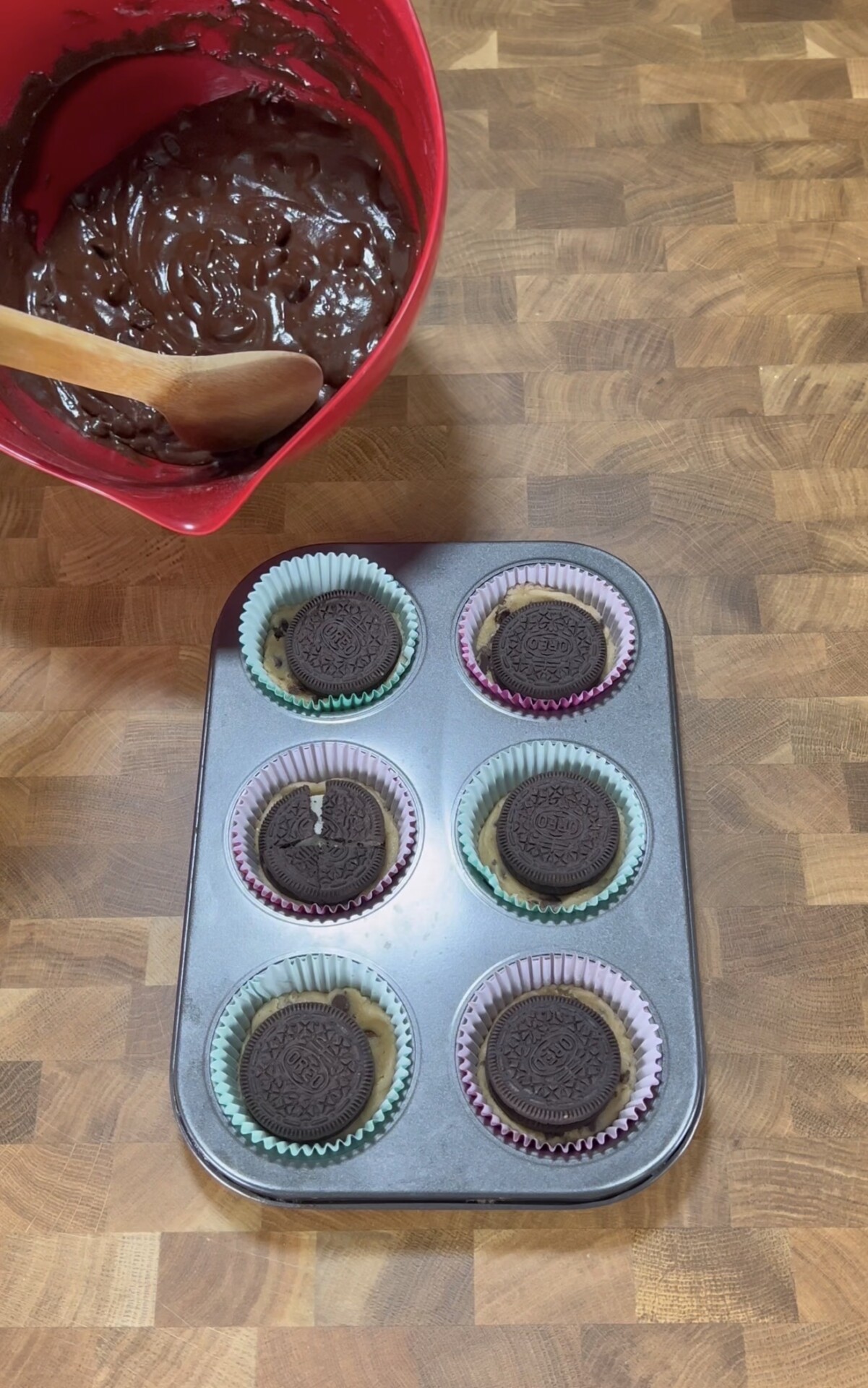 A muffin tin lined with pastel cupcake wrappers, each holding cookie dough and an Oreo cookie, sits on a wooden countertop next to a red bowl of chocolate batter with a wooden spoon.