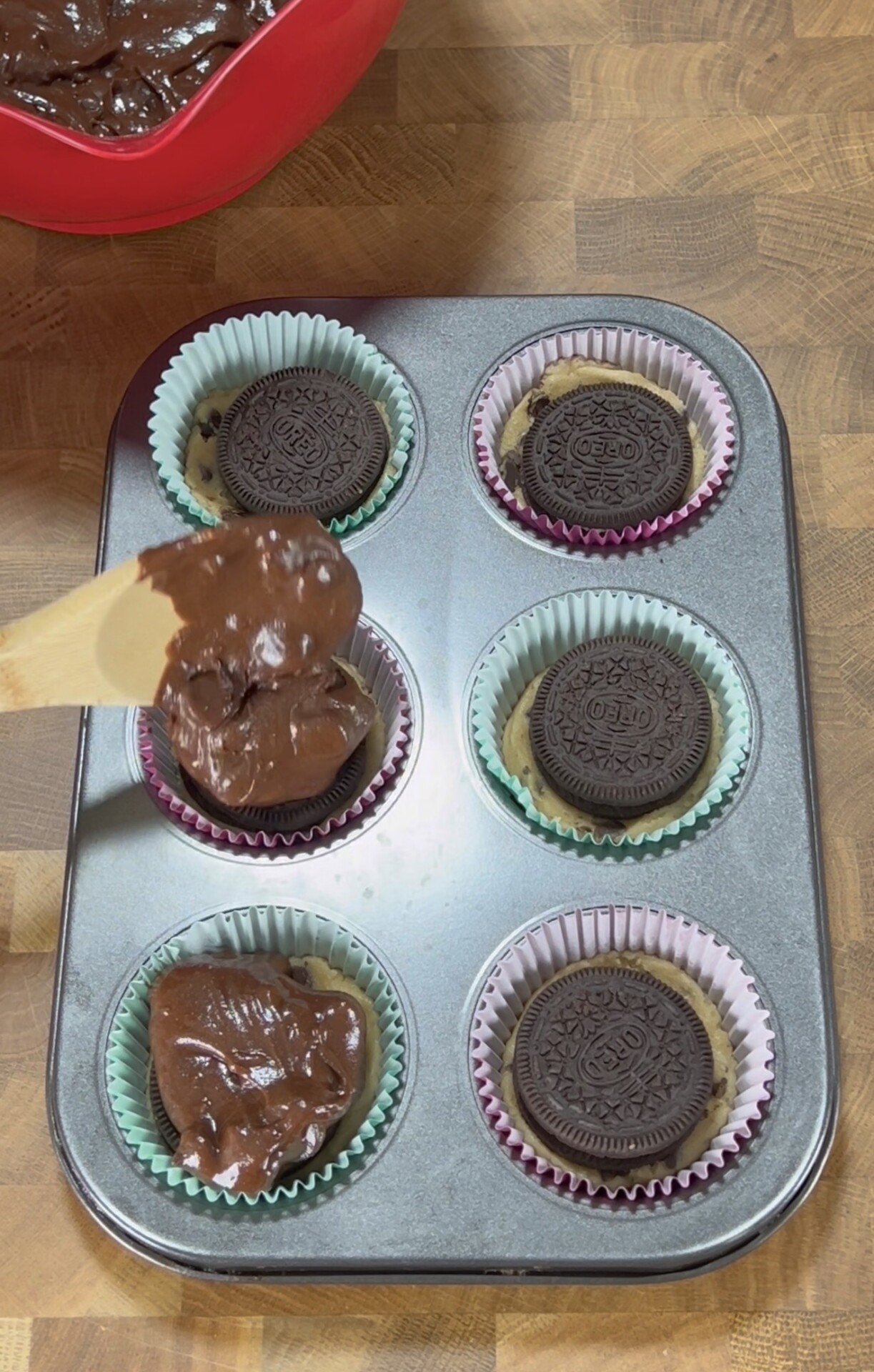 A muffin tray with six paper liners, each holding a cookie at the bottom. A hand is spooning chocolate batter over one cookie. A red bowl of more batter is visible in the top left corner.