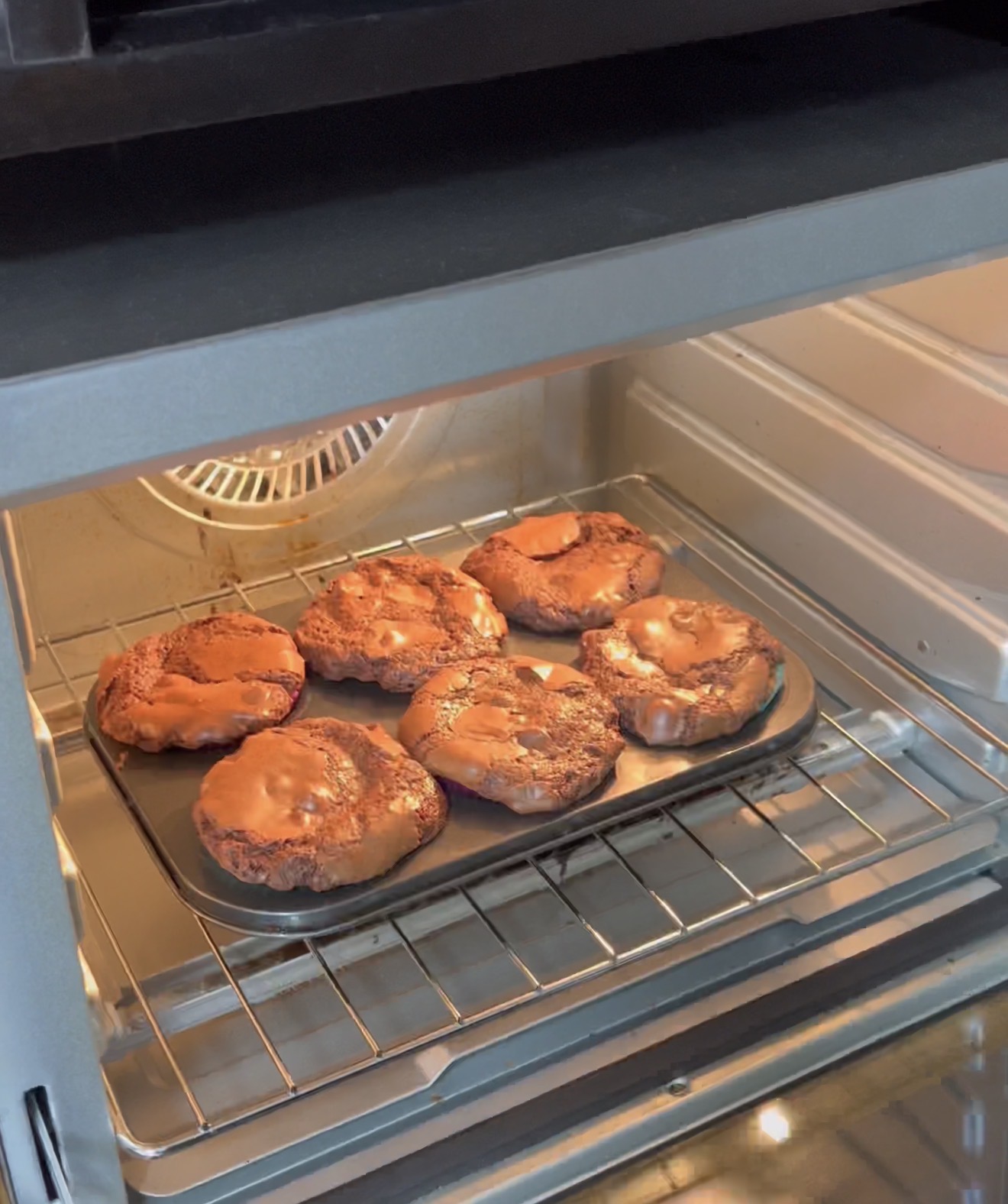 Six round chocolate cookies with melted caramel on top are baking on a metal tray inside an oven, with the oven door open and the oven light on.