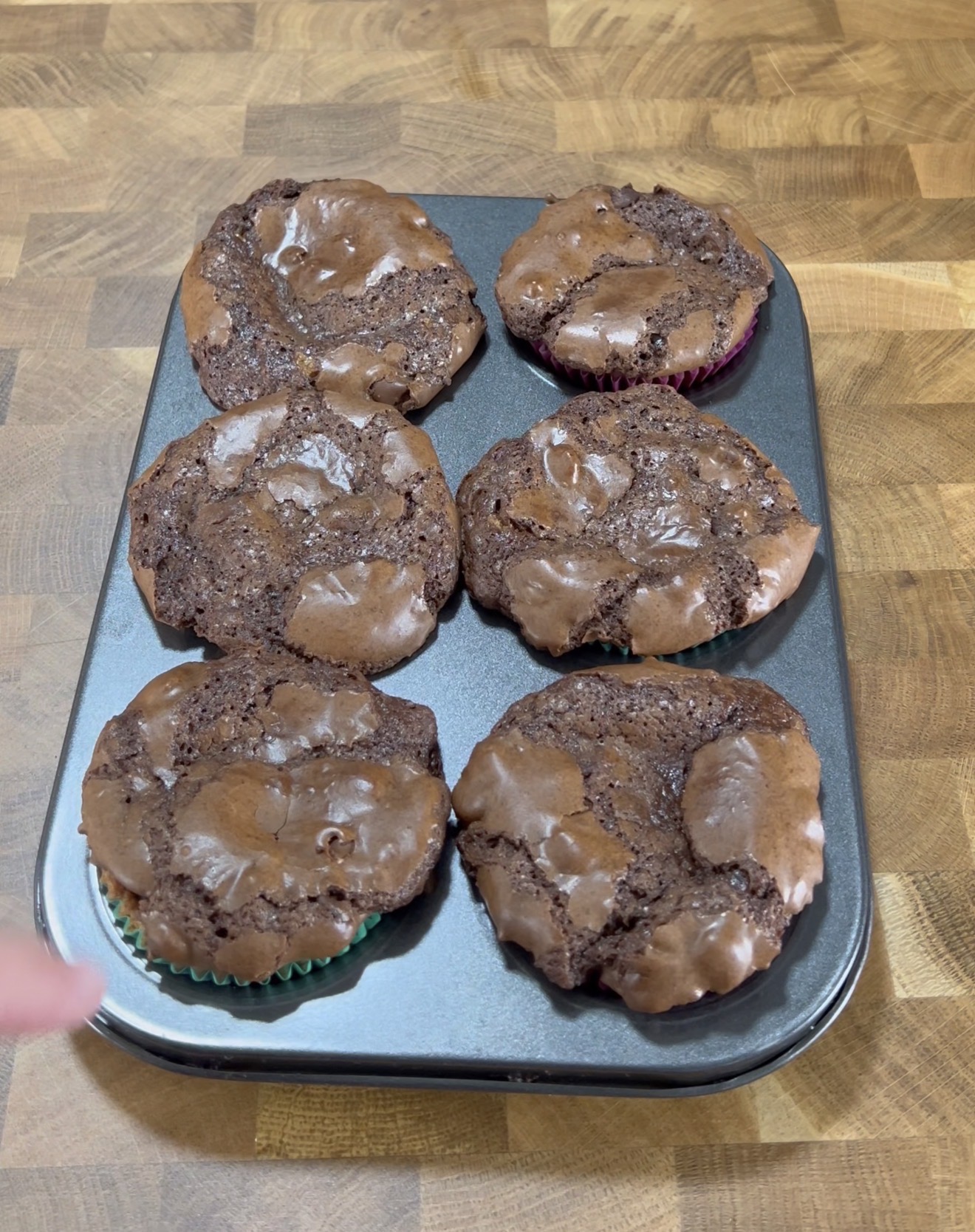 Five large, homemade chocolate muffins with cracked tops sit in a muffin tray on a wooden surface. The muffins have spread out and merged at the edges. A finger is visible at the bottom left of the image.