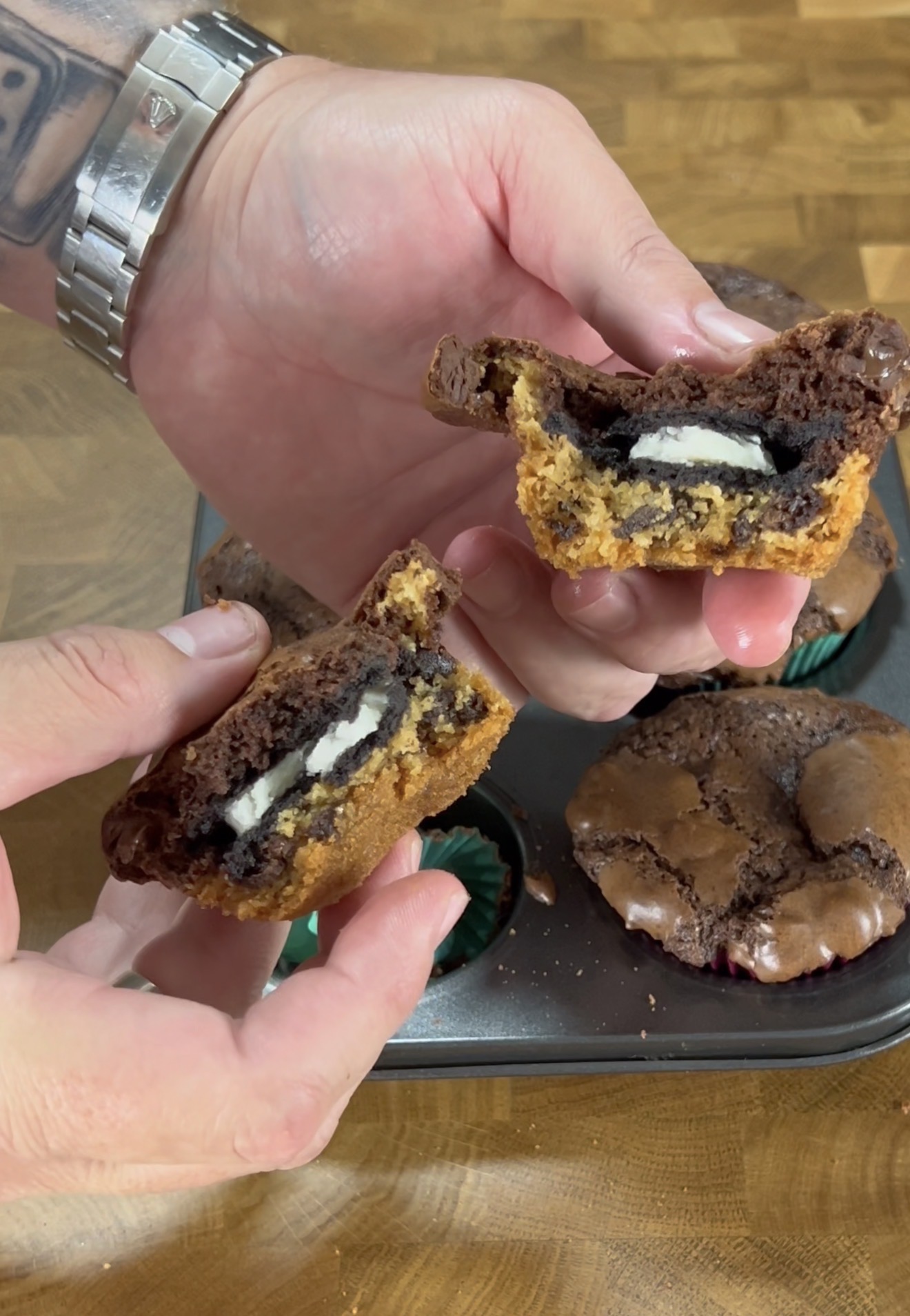 A person holds a split cookie cup showing layers of chocolate brownie, cookie dough, and an Oreo inside; more cookie cups are in a baking tin below.