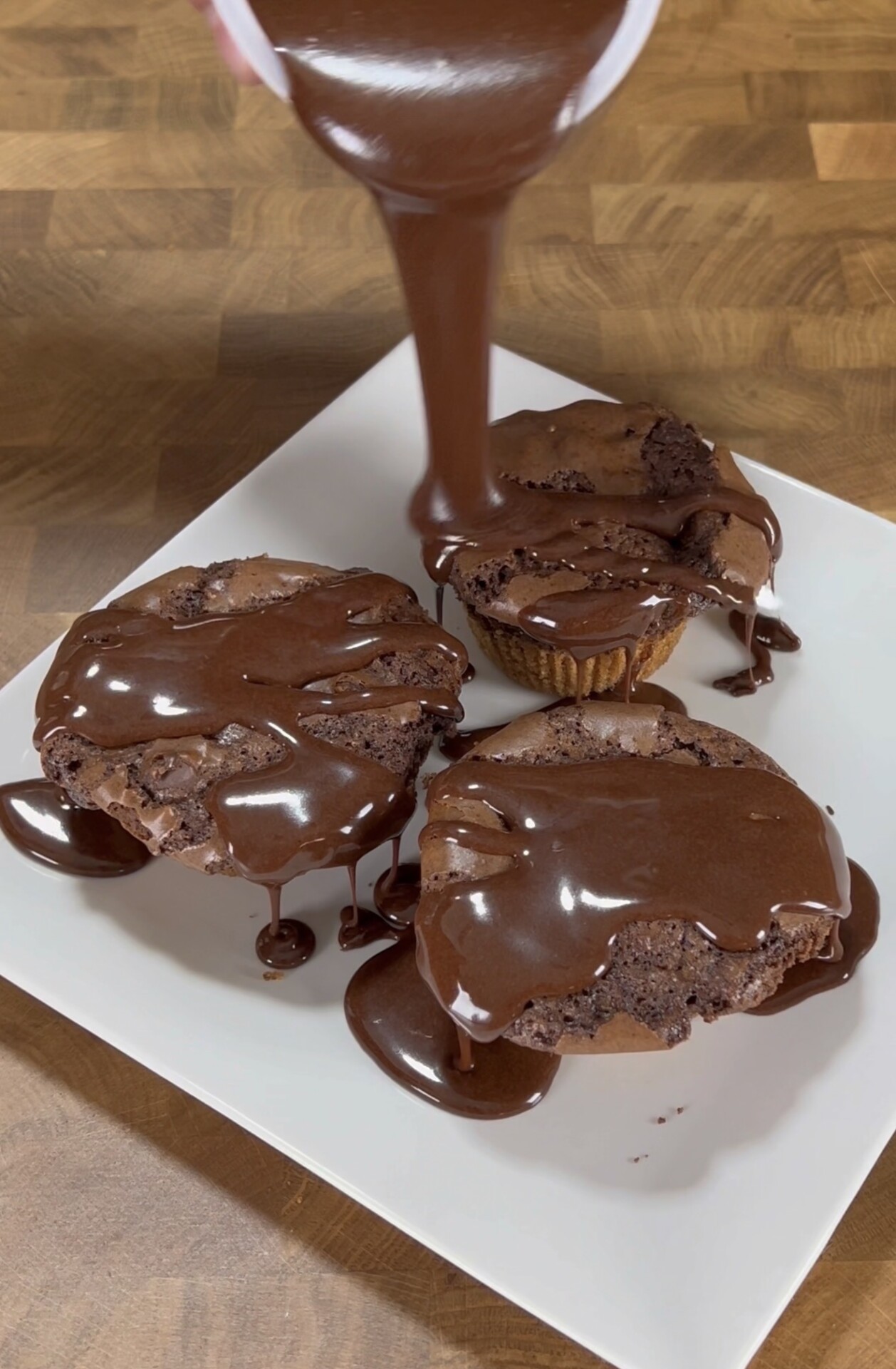 Three chocolate muffins on a white plate being generously drizzled with thick chocolate sauce, with some sauce pooling around the muffins on a wooden surface.
