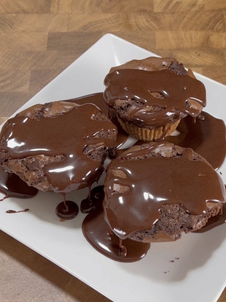 Three chocolate muffins are topped with a glossy layer of melted chocolate, displayed on a white square plate atop a wooden surface. The chocolate drips down the sides, creating a rich, indulgent appearance.