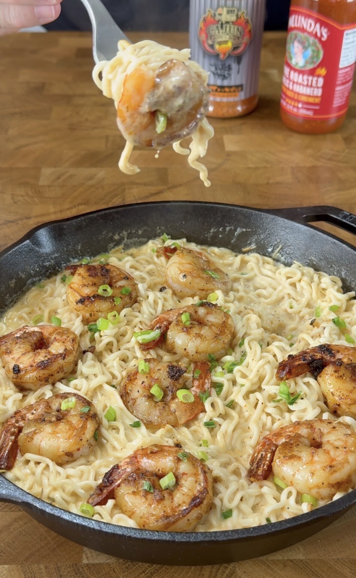 A skillet filled with creamy ramen noodles topped with grilled shrimp and chopped green onions. A hand holds a fork lifting a shrimp and some noodles. Two hot sauce bottles are visible in the background.