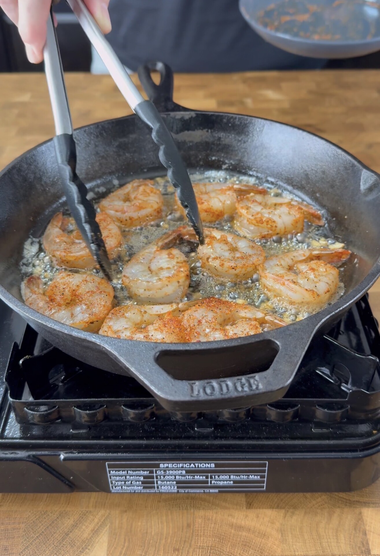 A person uses tongs to cook seasoned shrimp in a black cast iron skillet on a stovetop, with bubbling oil and bits of garlic visible in the pan.