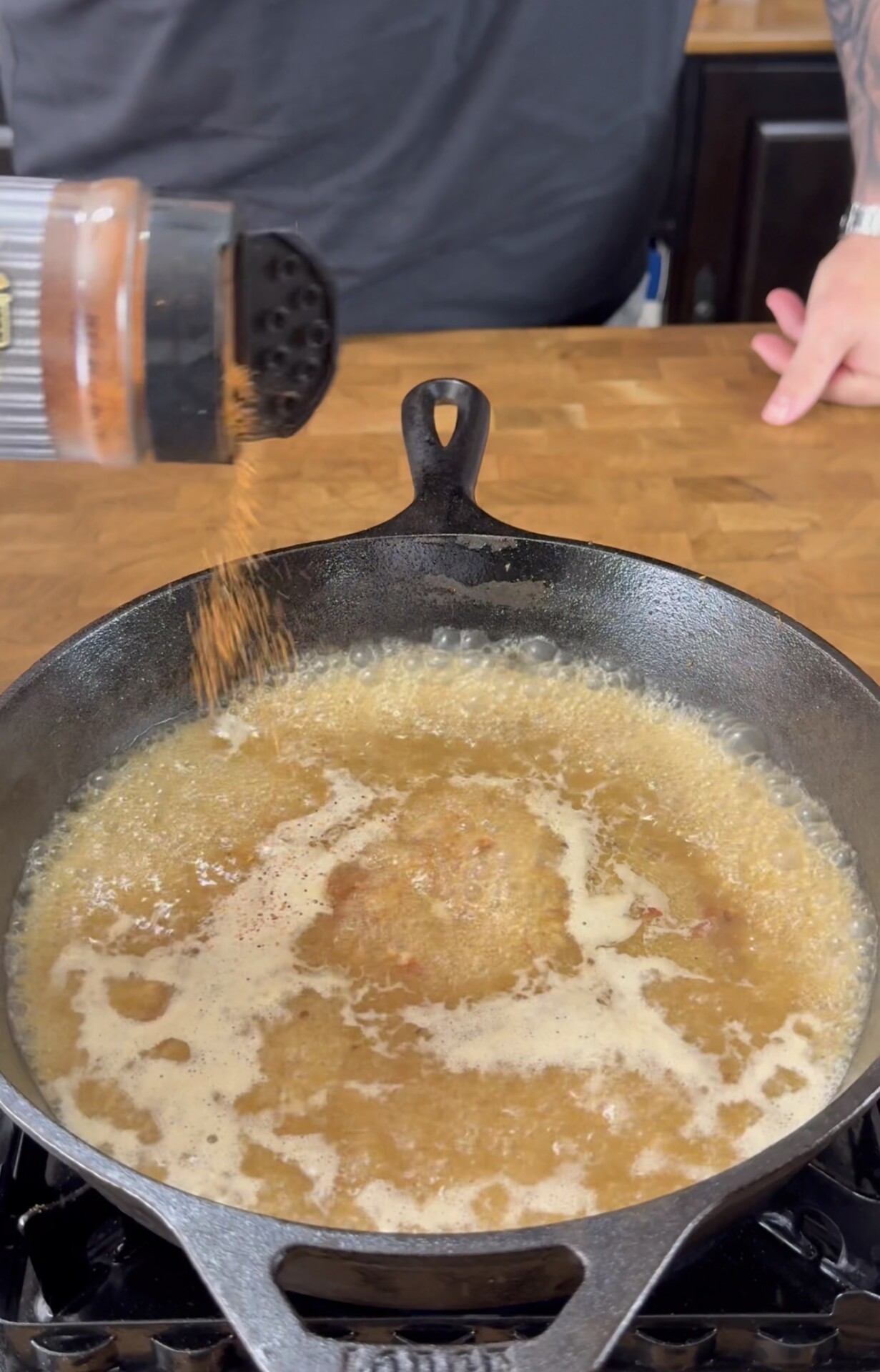 A person sprinkles seasoning from a container into a cast iron skillet filled with bubbling liquid on a stovetop, with a wooden countertop in the background.