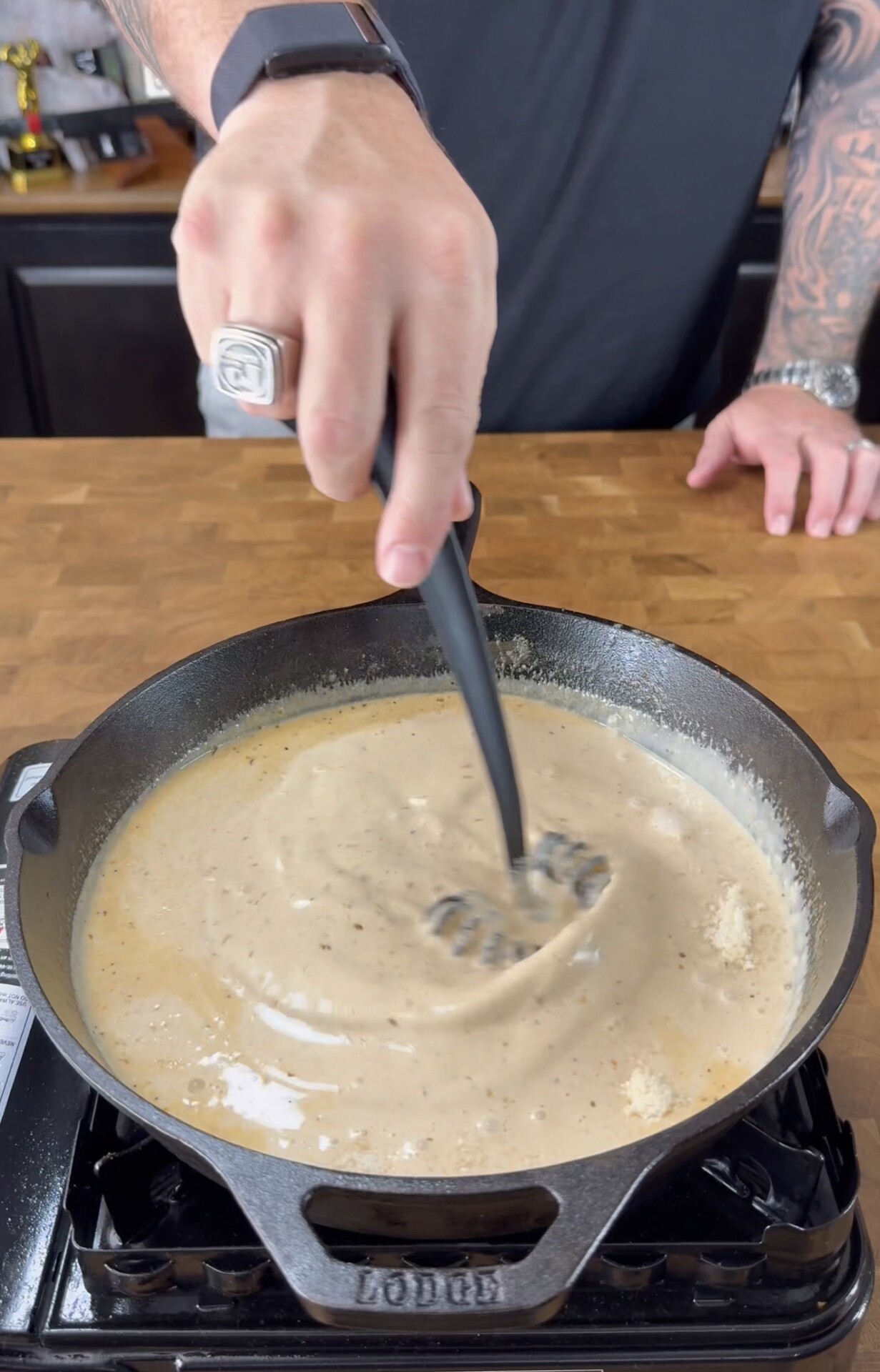 A person with tattooed arms stirs a thick, creamy sauce in a black cast iron skillet on a stovetop, using a black spatula. The scene is set on a wooden countertop.