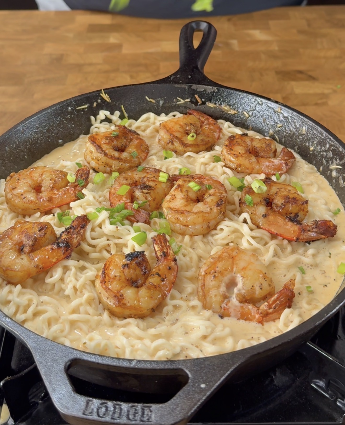A cast iron skillet filled with creamy noodles topped with grilled shrimp and garnished with chopped green onions, sitting on a wooden surface.