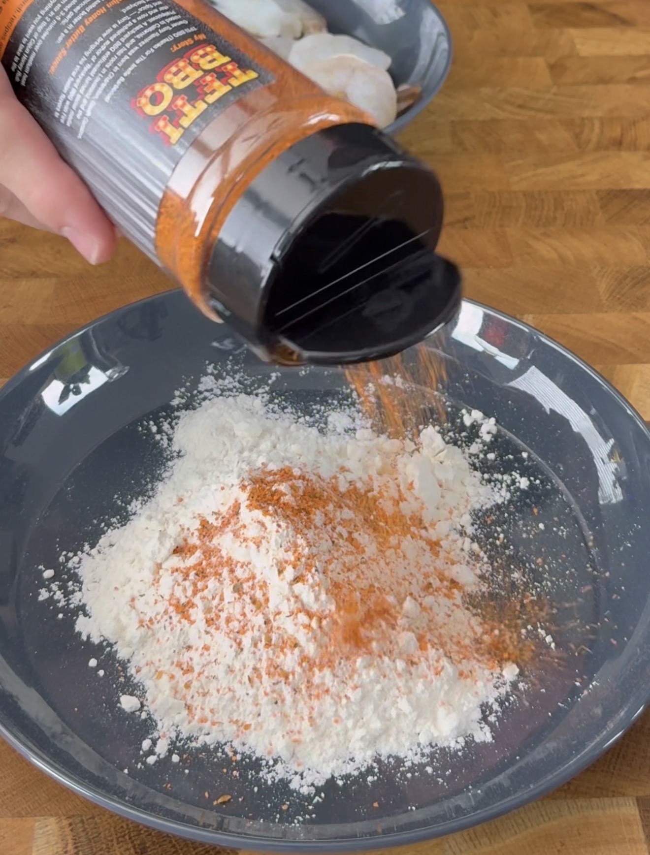 A hand sprinkles orange spice from a container labeled BBQ onto a gray plate with a mound of white flour, on a wooden countertop. Another plate with raw shrimp is in the background.