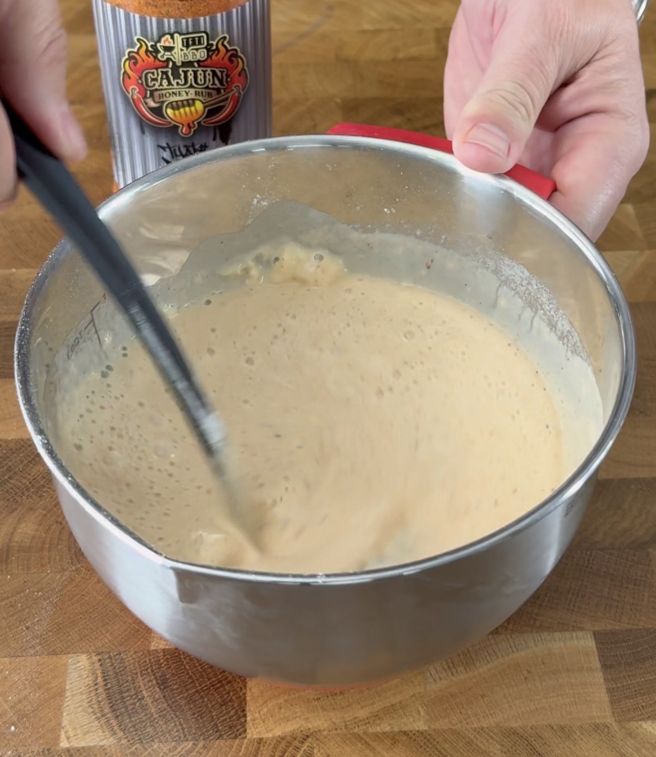 A person stirs a light brown batter in a metal mixing bowl with a black spatula. A can labeled Cajun Injector is visible in the background on a wooden surface.