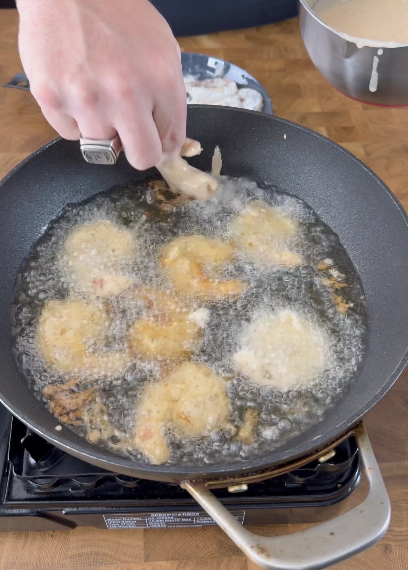 A hand lowers a battered shrimp into a frying pan filled with hot oil, where several other shrimps are being deep-fried on a stovetop. A bowl with more batter is visible in the background.