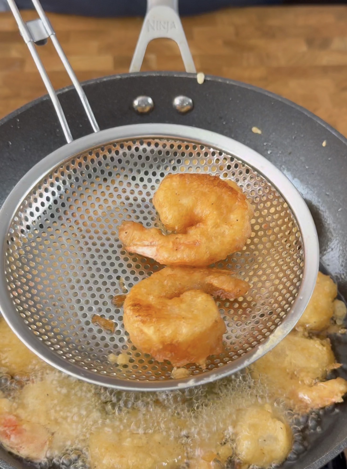 A metal strainer holds two golden-brown fried shrimp above a pan of hot oil, with more shrimp frying below. The background features a wooden surface.