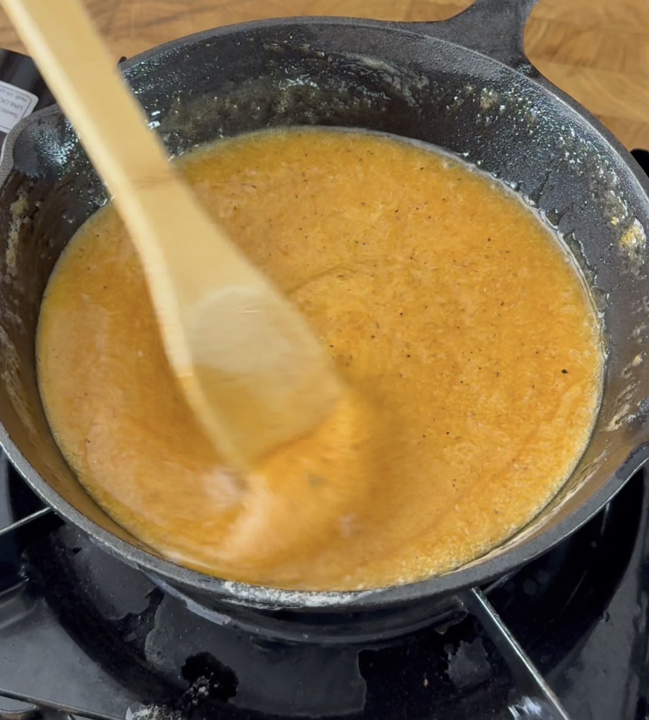 A wooden spoon stirs a creamy orange sauce in a black cast iron pan on a stovetop, with a wooden countertop visible in the background.