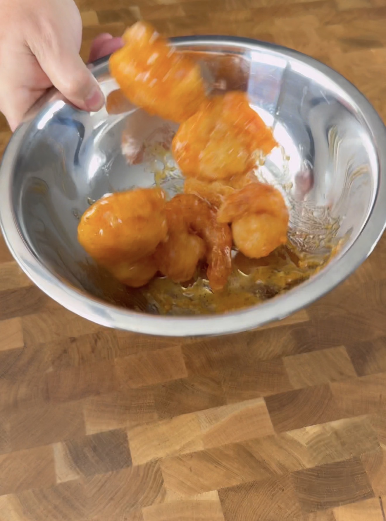 A hand tossing crispy, orange-sauced shrimp in a stainless steel bowl over a wooden surface.